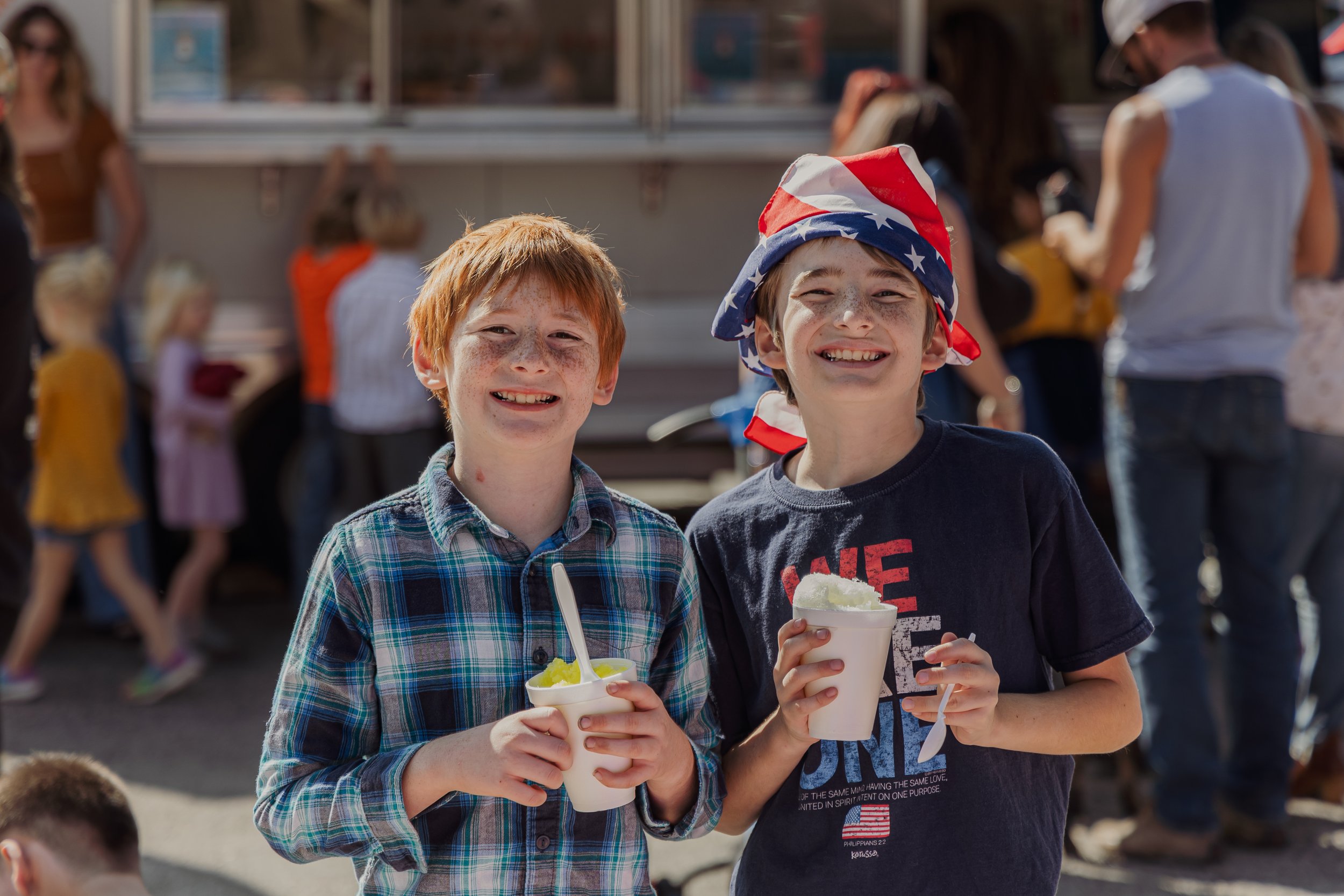 Two boys smiling and holding cups of ice cream at an outdoor event, with a crowd of people in the background.