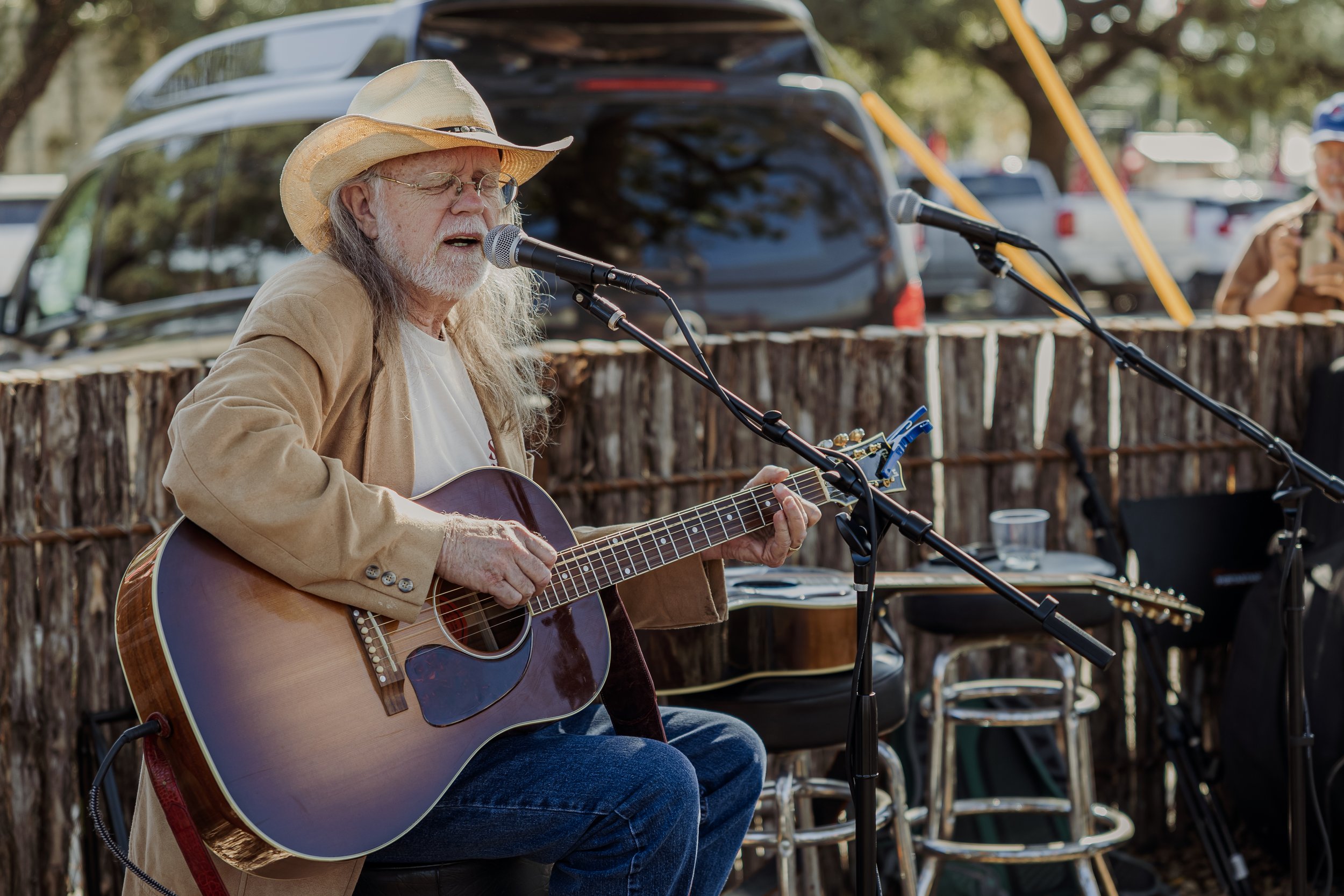 An elderly man with long gray hair, a long beard, wearing a wide-brimmed hat, glasses, a beige jacket, and a white t-shirt, playing an acoustic guitar and singing into a microphone outdoors. There is a wooden fence behind him, a black car, and a pers