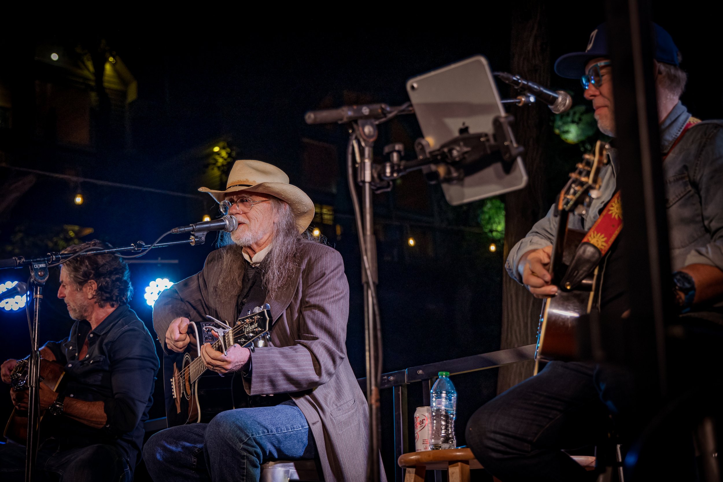 Three men playing acoustic guitars on stage during a live performance at night, with one wearing a cowboy hat and glasses, and two others with the right side in focus, microphones and stage lighting visible.