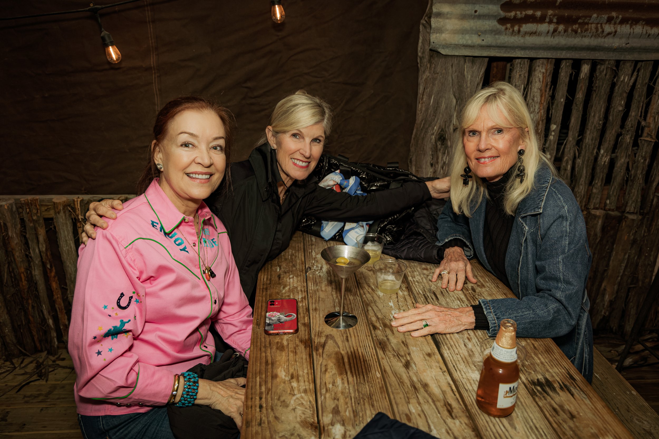 Three women sitting at a rustic wooden table in a dimly lit setting, smiling at the camera. The woman on the left is wearing a pink shirt with embroidery, the woman in the middle is in black, and the woman on the right is in a denim jacket. There are