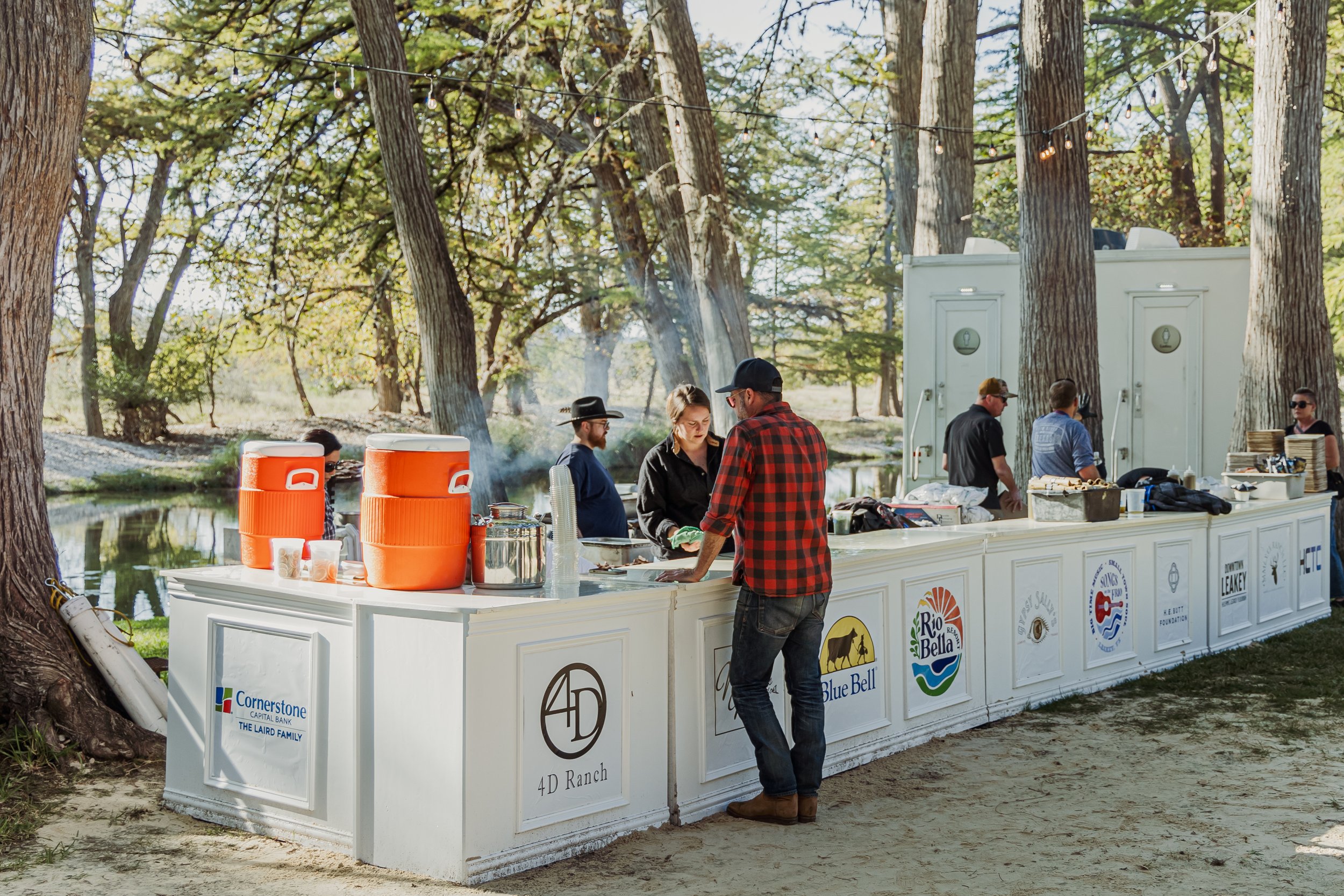 An outdoor event with a long white bar setup by a pond, serving drinks to people. There are large orange coolers on the bar, and several people are standing or working behind it. The background features trees and string lights hanging above.