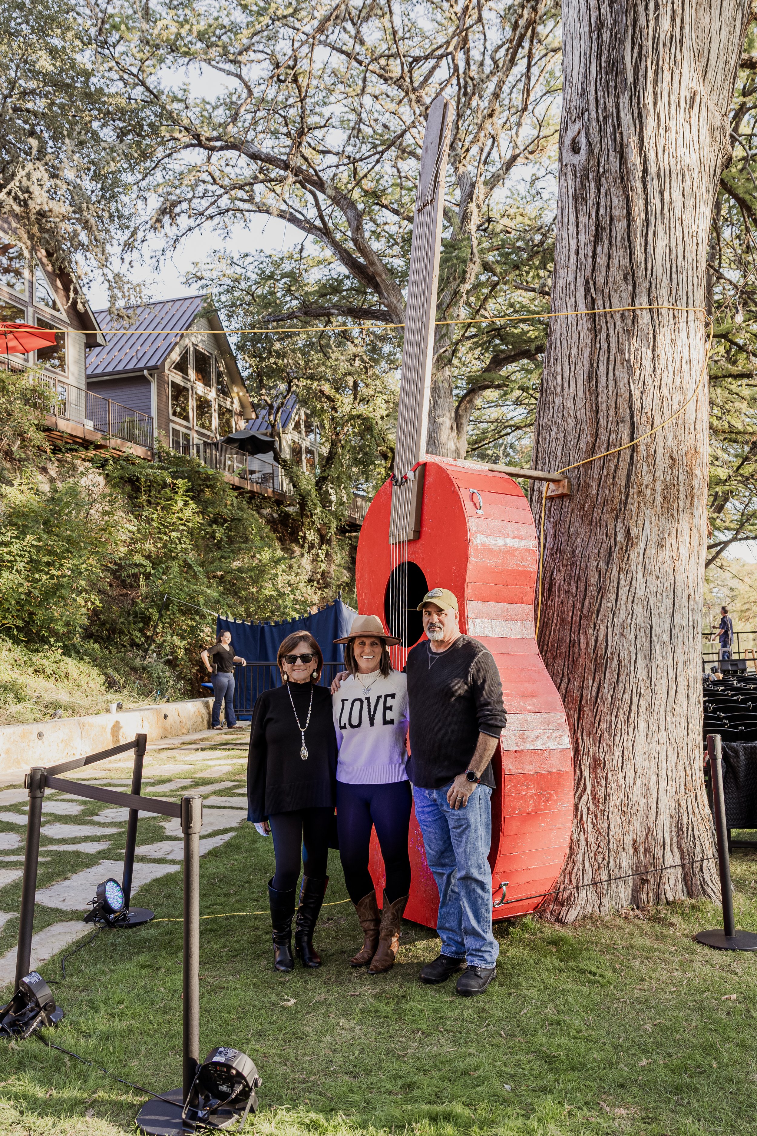 Three people standing in front of a large painted red and gray guitar-shaped sculpture next to a tree outdoors, with a house with large windows and a balcony on a hillside in the background.