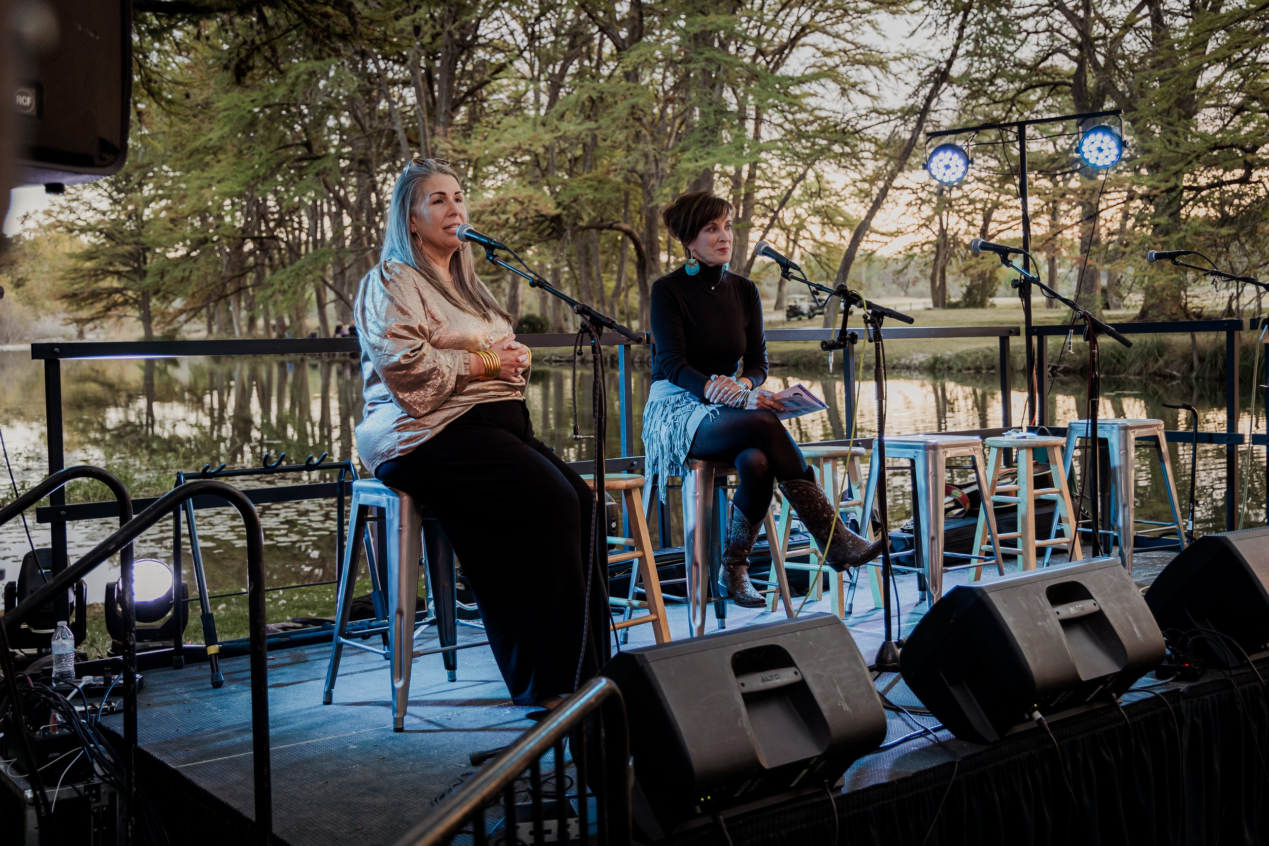 Two women sitting on stools on an outdoor stage by a lake, speaking into microphones, with trees and water in the background during sunset.