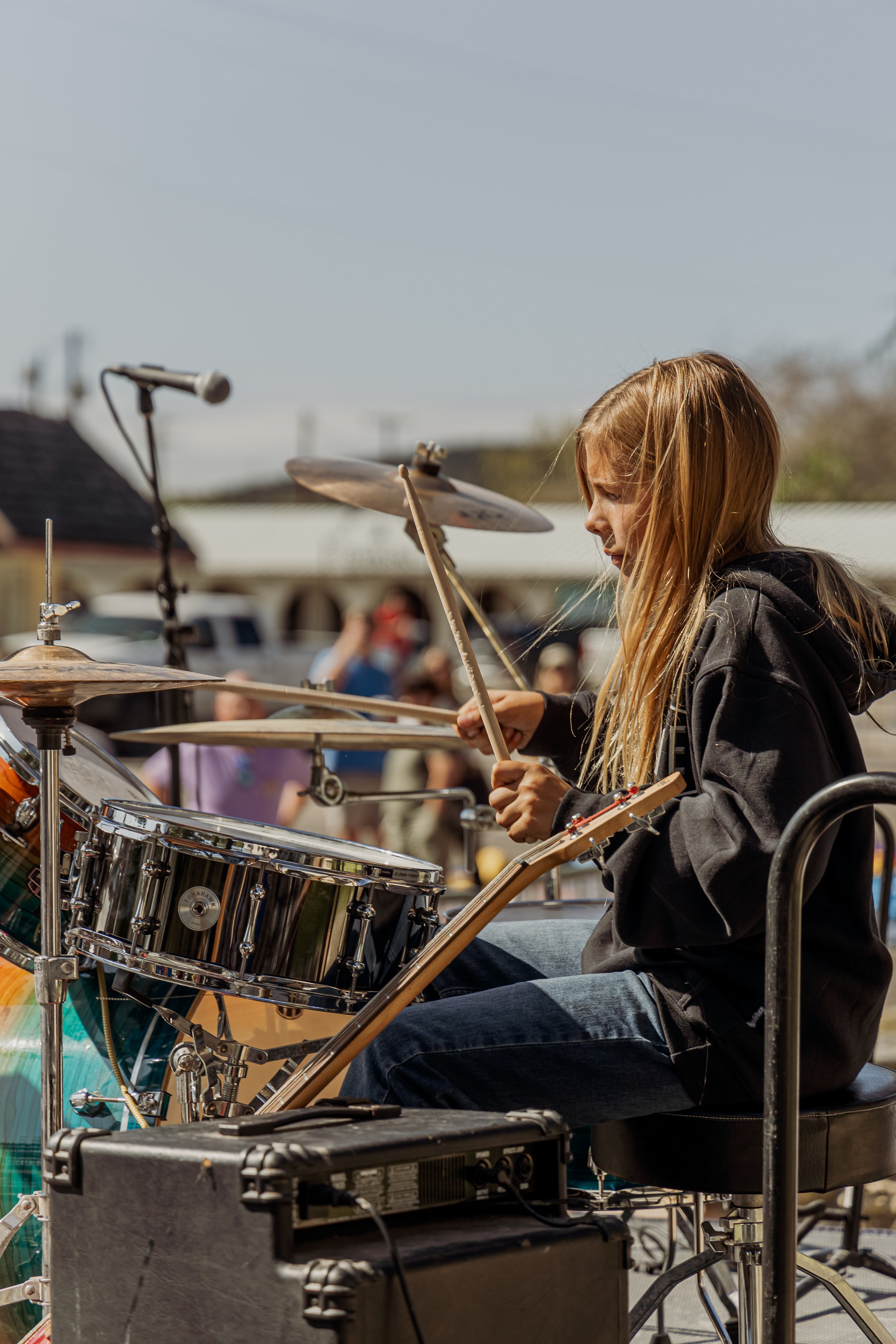 A young girl playing drums on an outdoor stage during daytime, with a crowd and houses in the background.