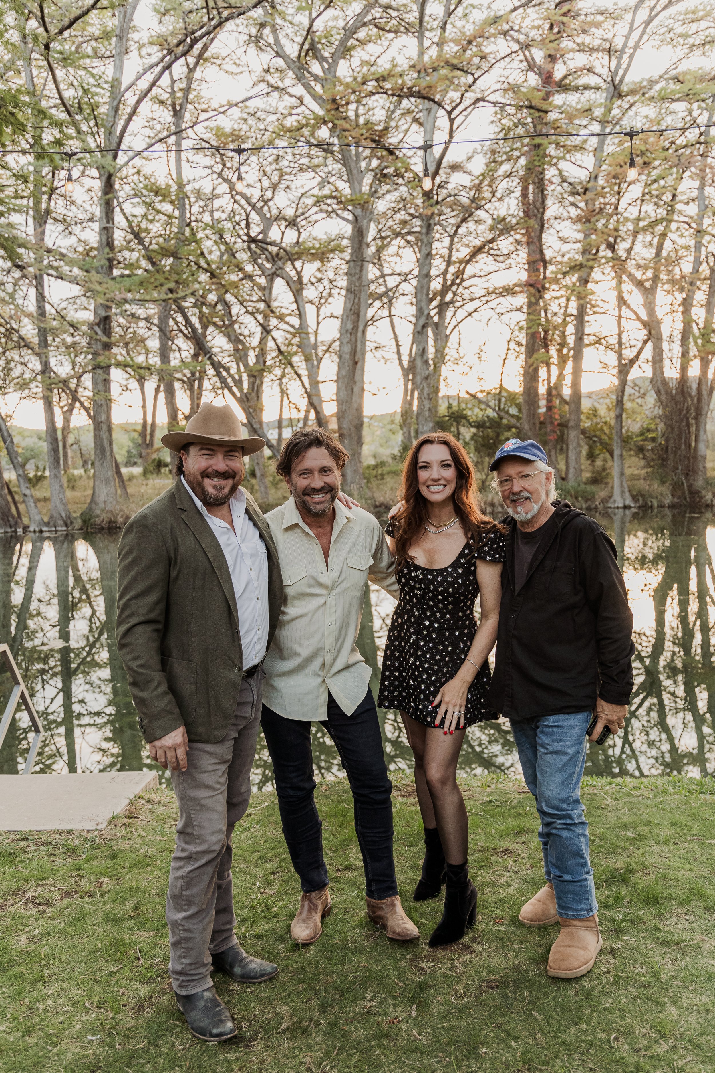 Four people standing outdoors by a lake, smiling at the camera during sunset. Trees line the background and string lights are hanging overhead.