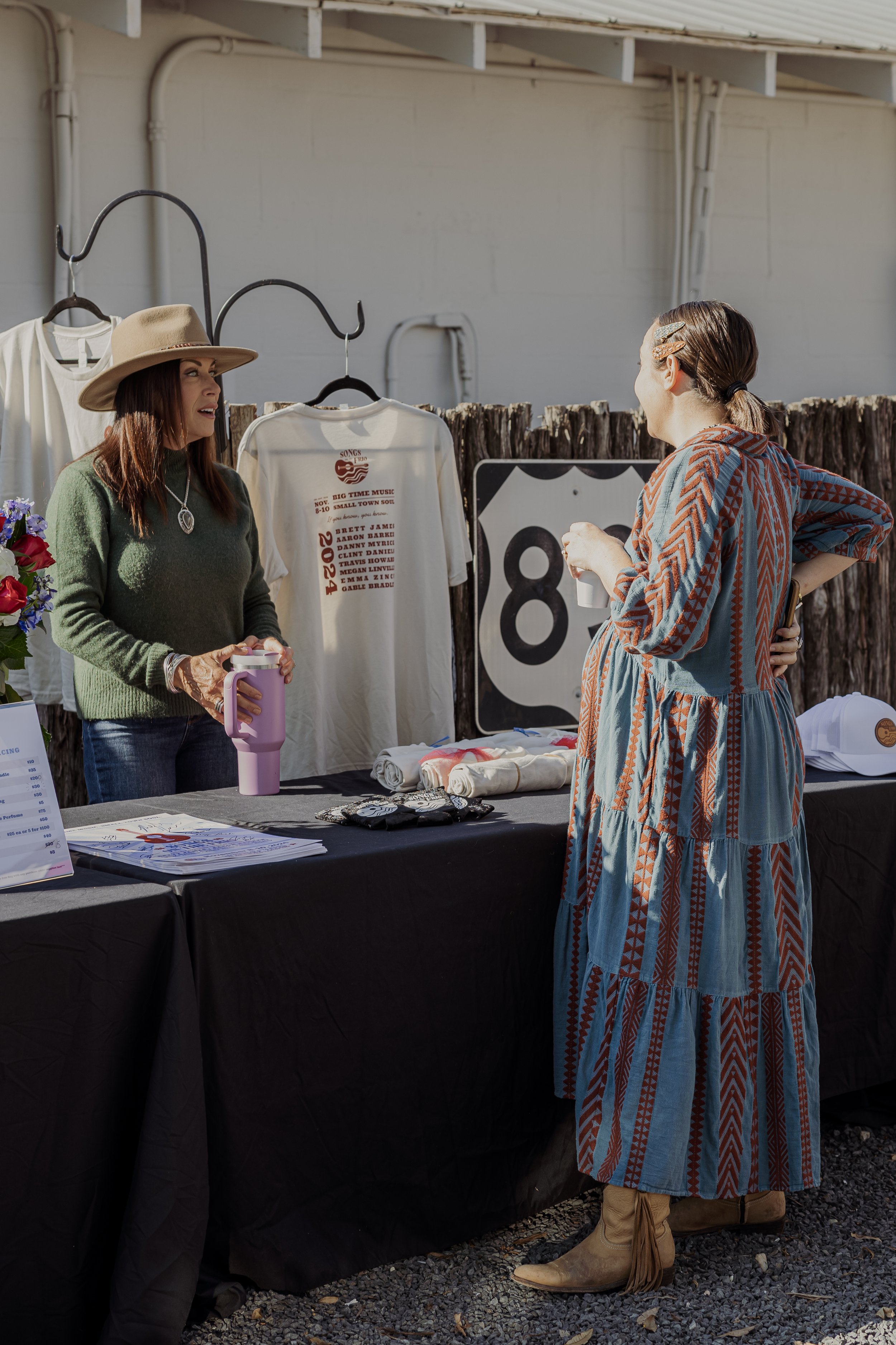 Two women talking at an outdoor merchandise booth with T-shirts, hats, and other items, set against a wooden fence and a road sign.