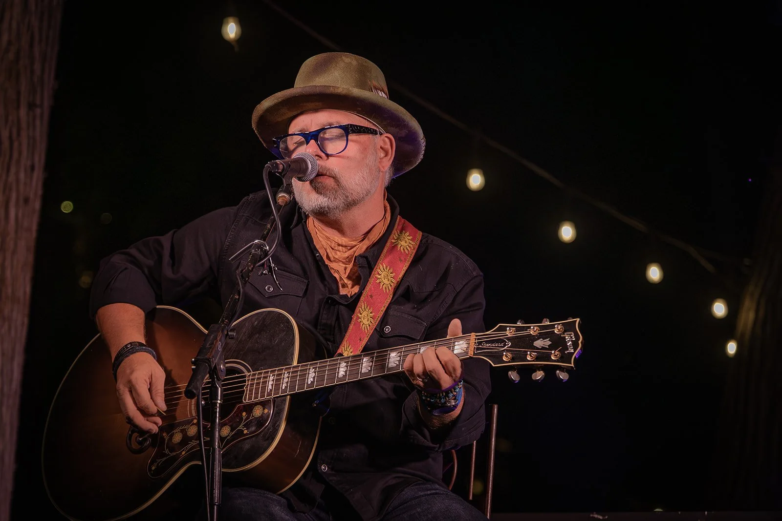 A man with glasses, wearing a fedora and black shirt, plays an acoustic guitar and sings into a microphone on an outdoor stage at night, with string lights above.