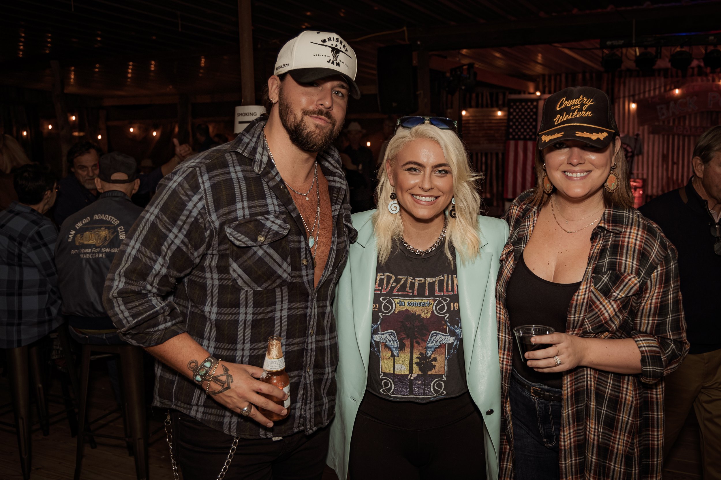 Three people smiling at a bar or pub, dressed in casual, country or western style clothing, with American flags and rustic decor in the background.