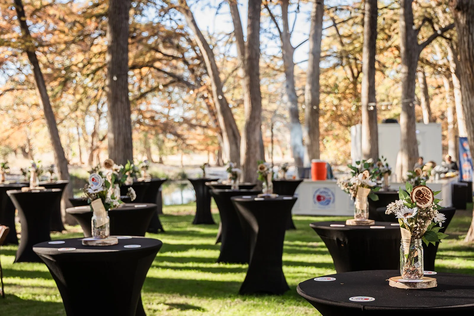 Outdoor event space with tall trees, with black cocktail tables decorated with glass jars filled with flowers and foliage, placed on wooden slices.