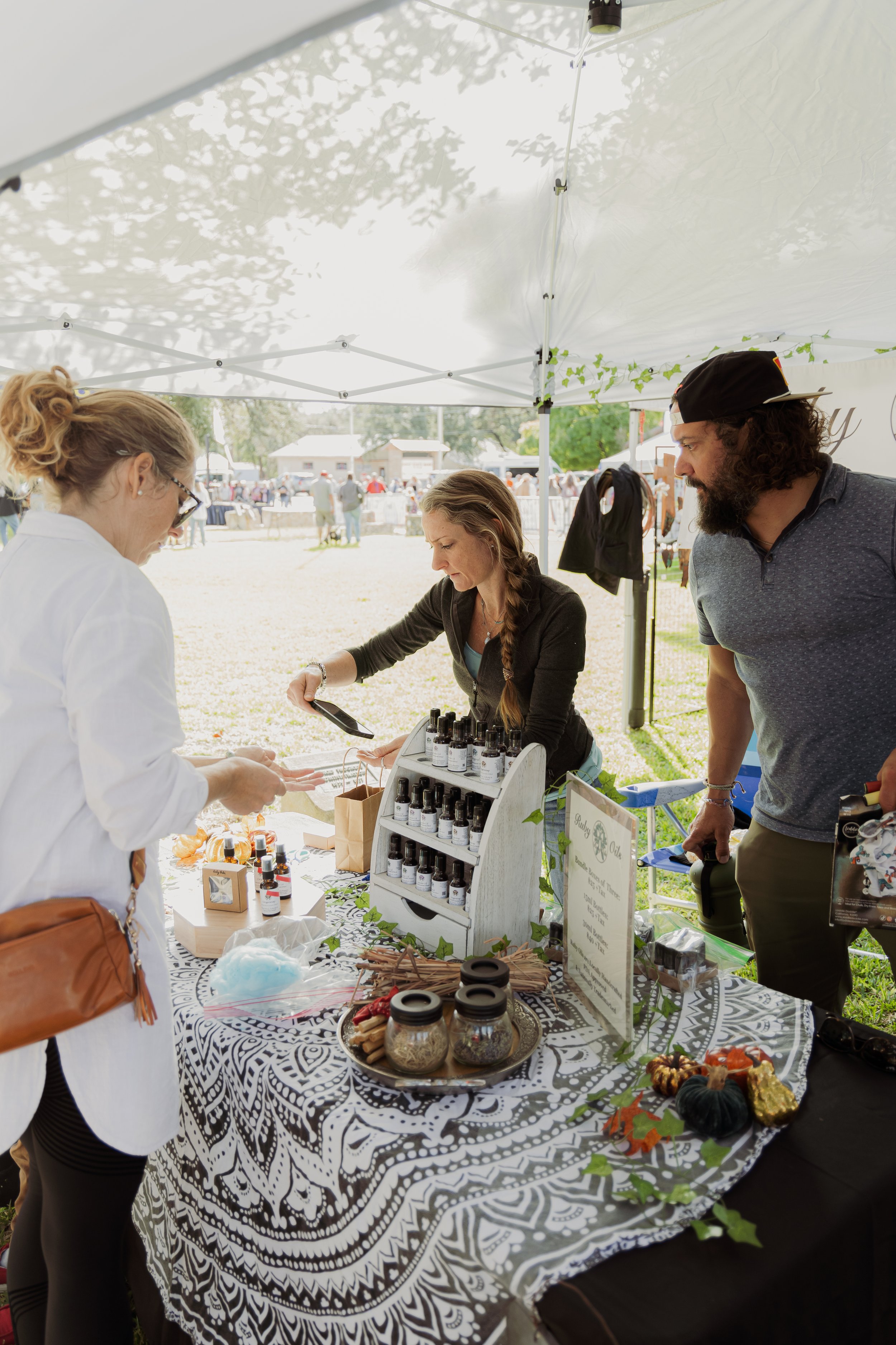 People at a vendor booth selling essential oils and handmade crafts at an outdoor market.