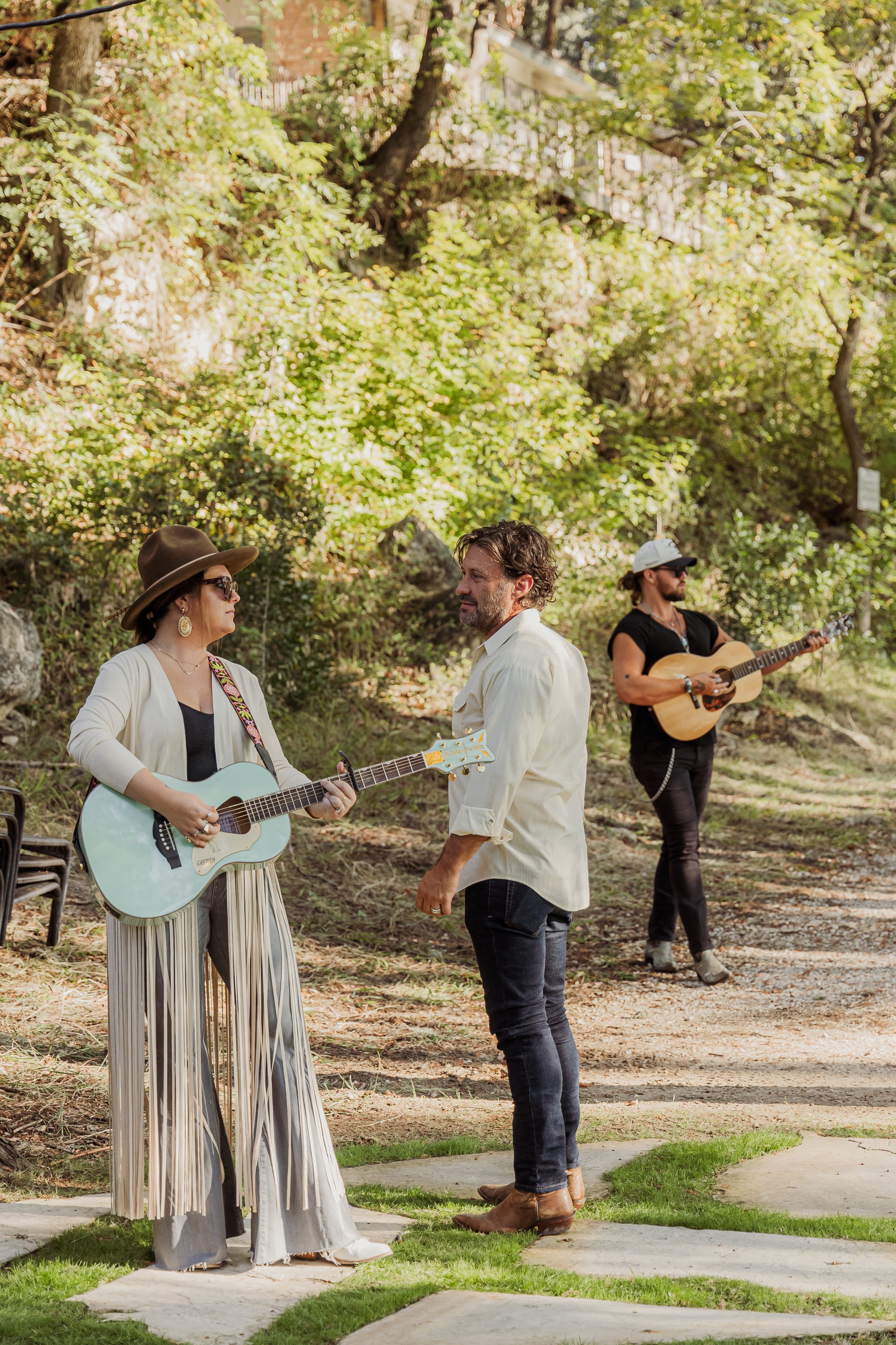Three musicians performing outdoors in a park-like setting with trees and greenery. A woman in a wide-brimmed hat and sunglasses plays a light blue guitar, a man in a light shirt and jeans appears to be singing or talking, and another man in a white