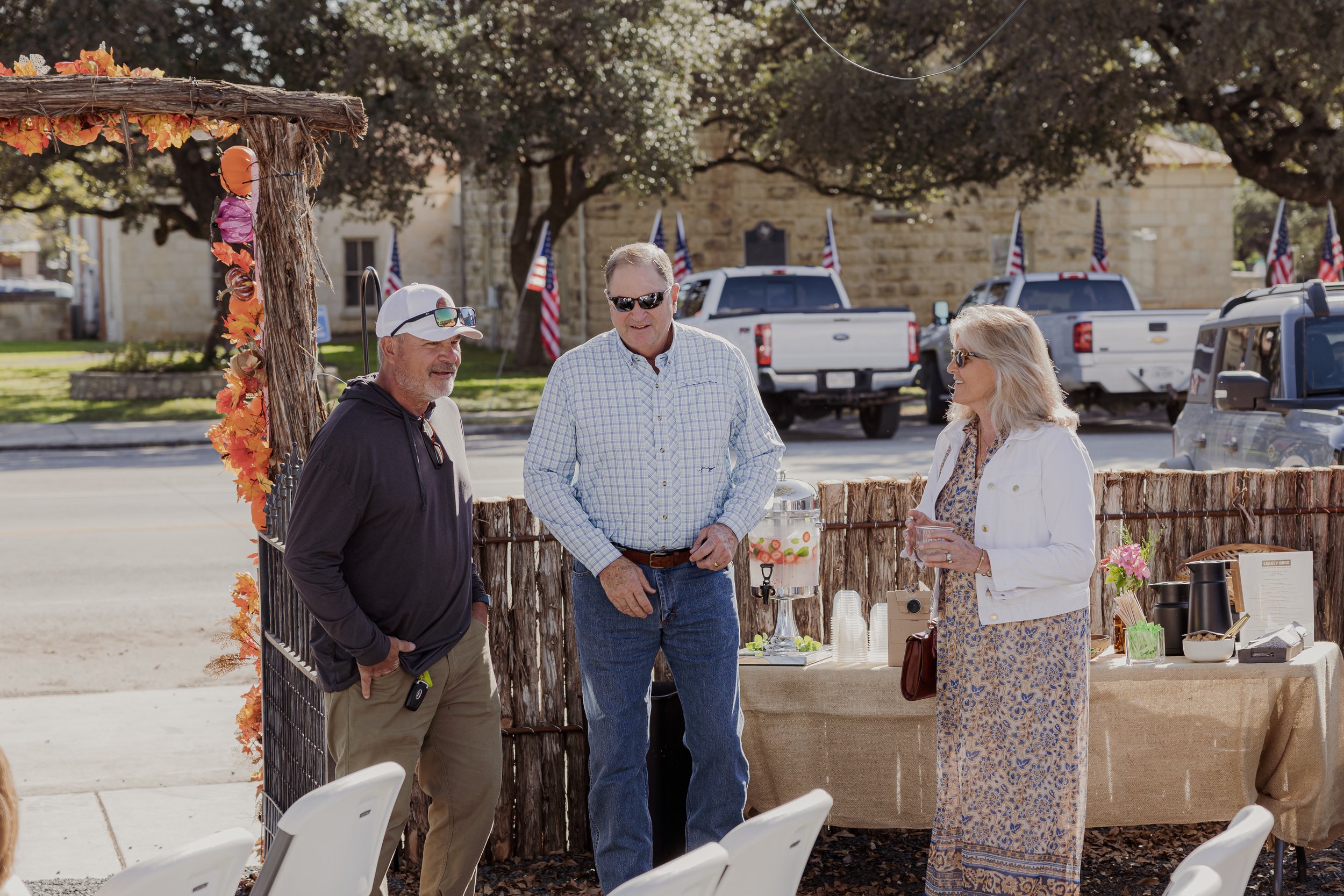 Three adults, two men and one woman, standing and talking at an outdoor event with a table behind them, decorated archway, and American flags in the background.