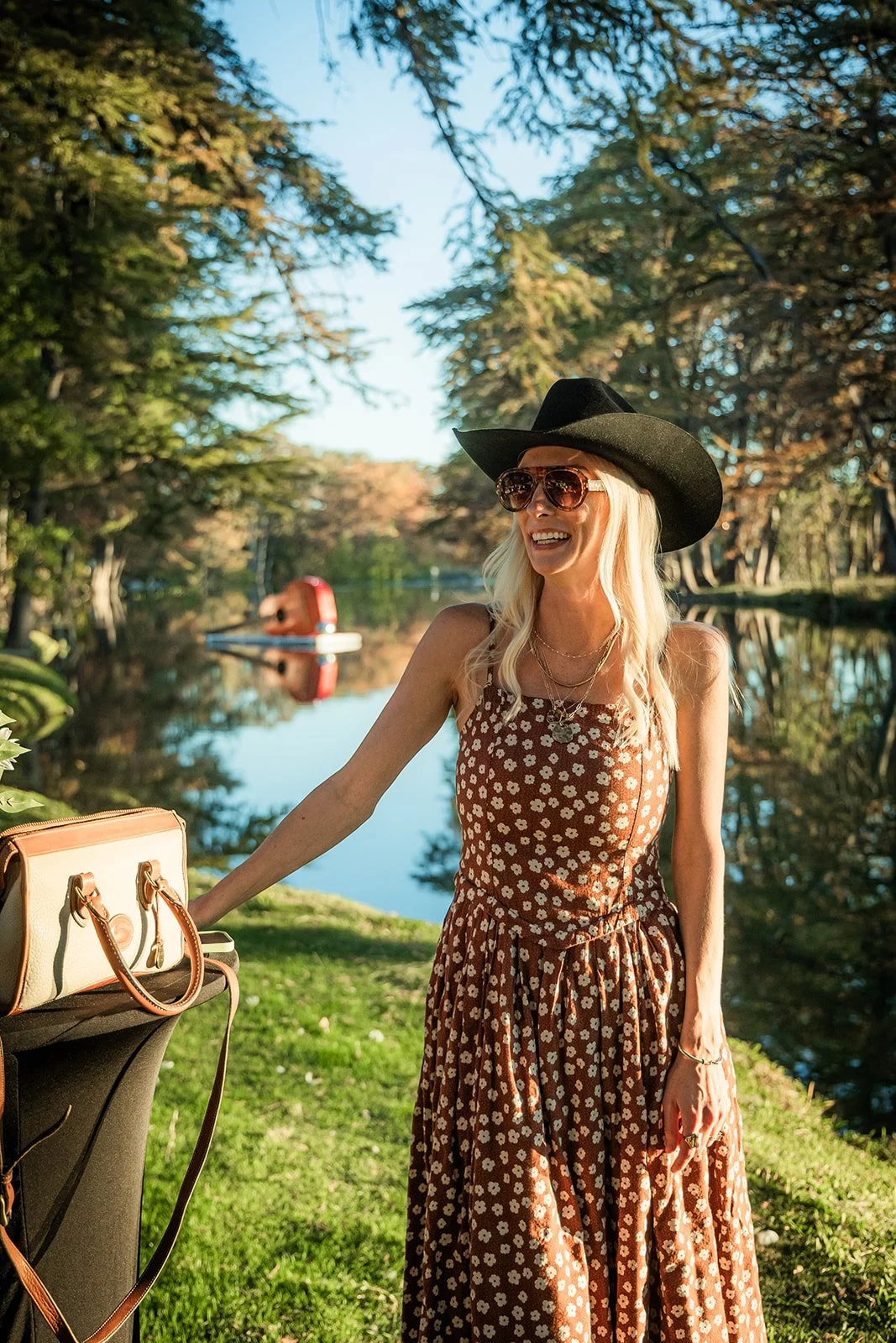 A woman in a long brown floral dress, wearing a large black hat and sunglasses, is standing outdoors next to a table with a bag, smiling in front of a peaceful lake surrounded by trees, with a red boat floating on the water.