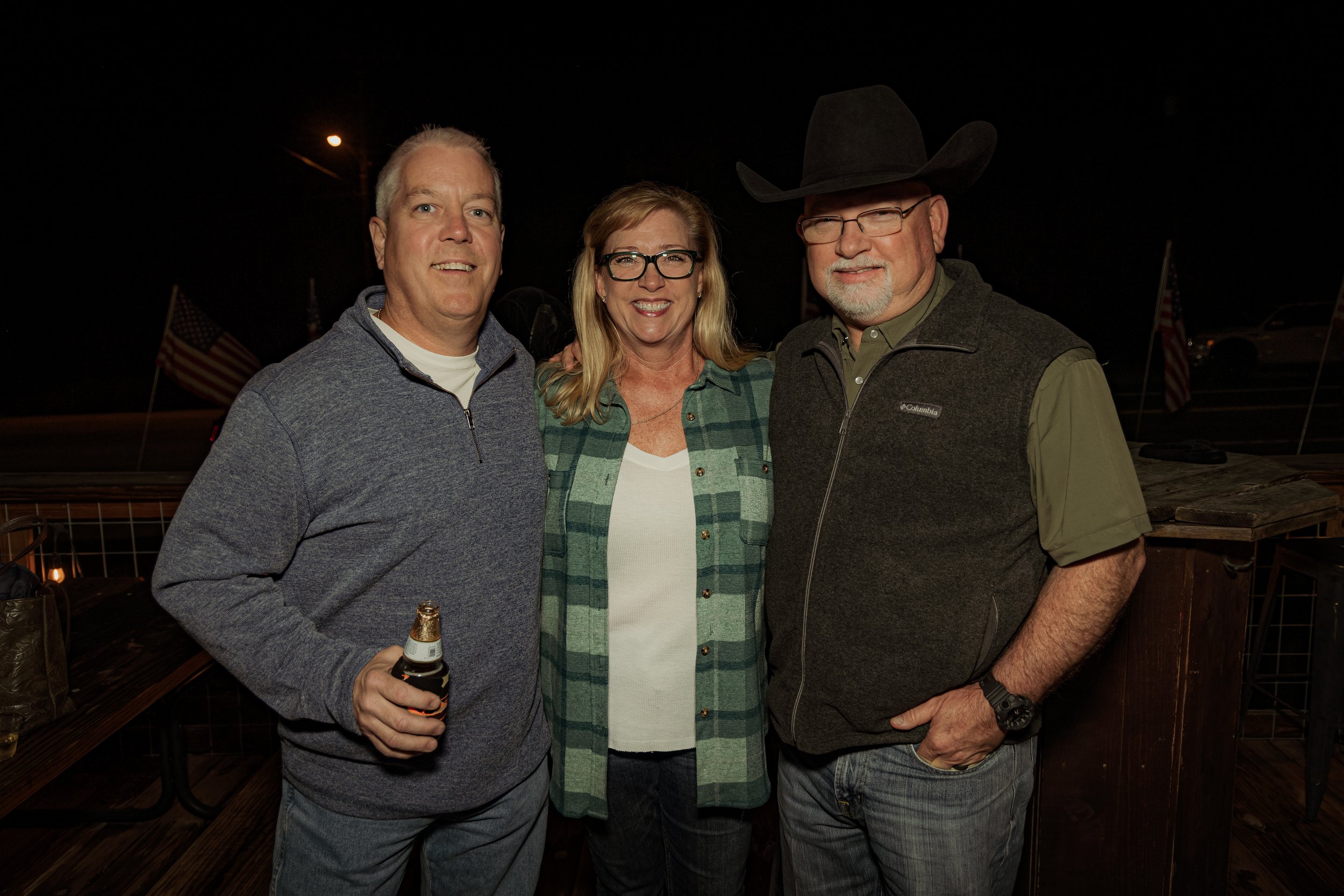 Three adults standing outdoors at night, smiling, with American flags and tents in the background