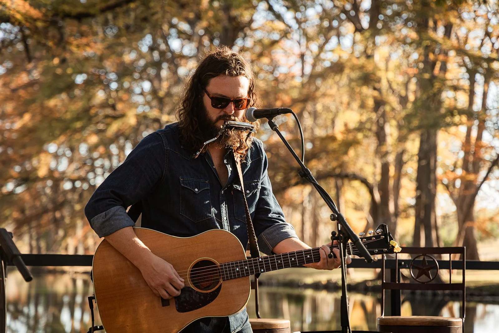 A man with long curly hair, beard, wearing sunglasses and a denim shirt, playing an acoustic guitar and singing into a microphone outdoors during autumn.