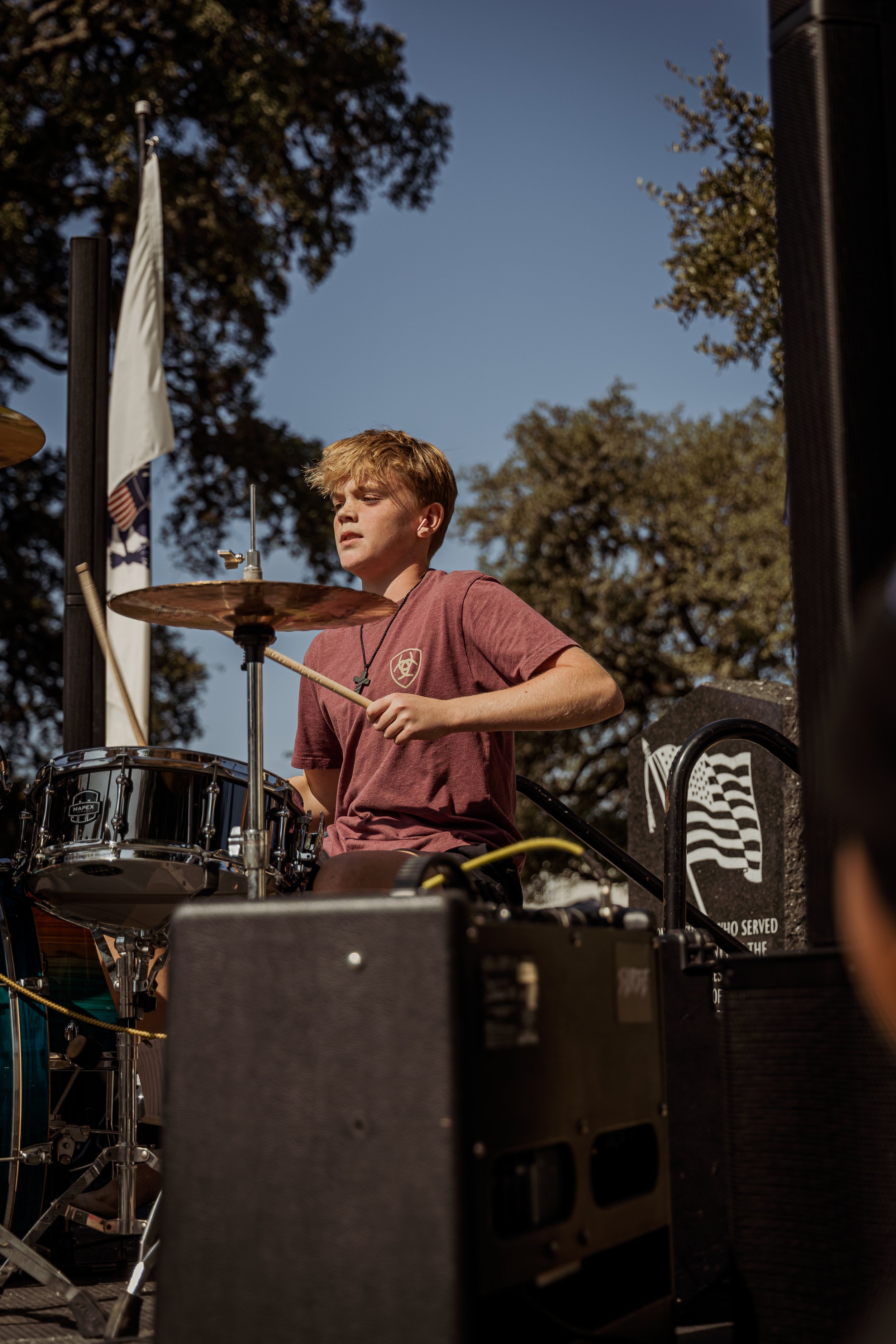 A young boy playing drums outdoors on a sunny day, with large trees and flags in the background.