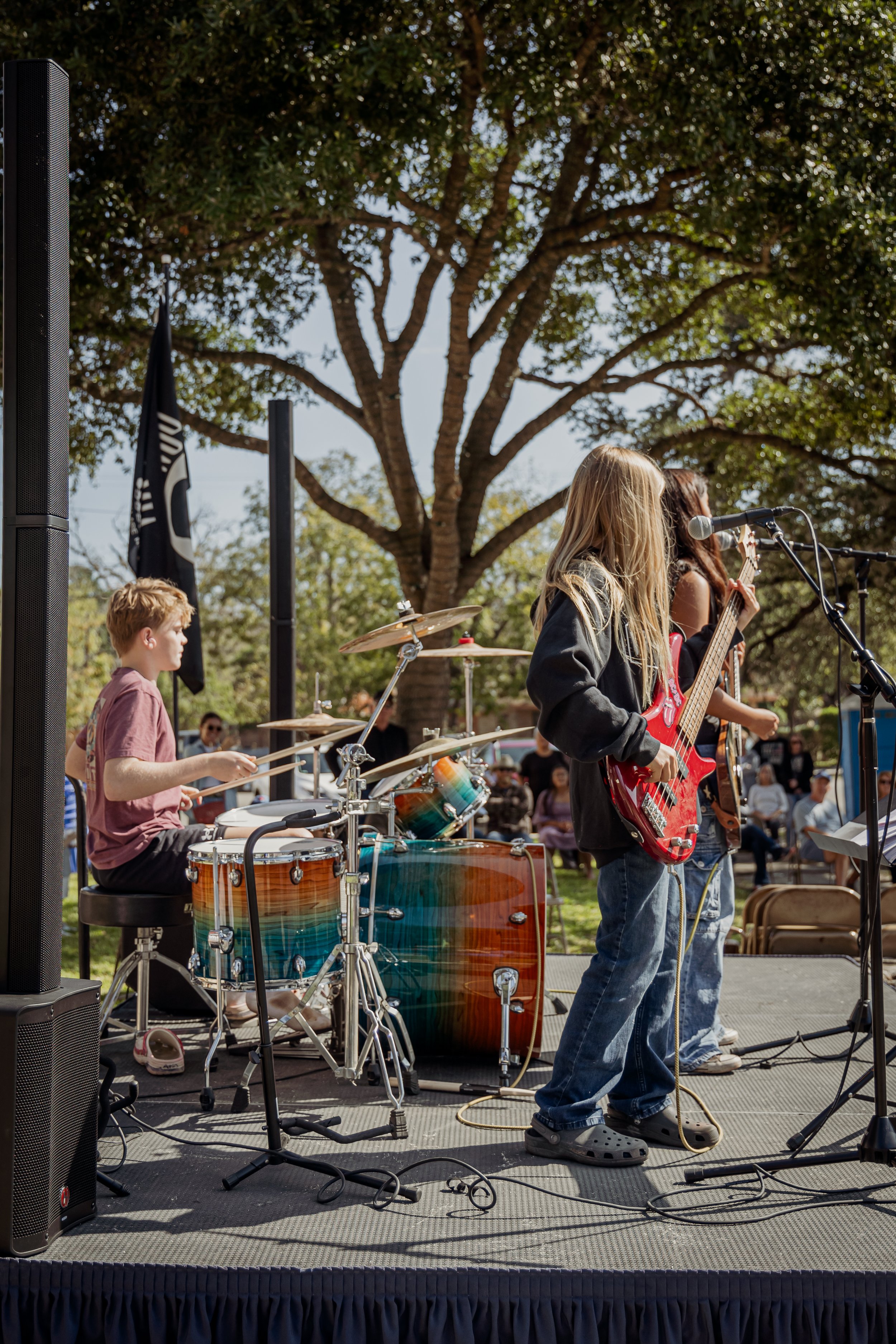 Children performing in a band outdoors, with a boy playing drums and girls playing guitar, in front of a large tree and audience.