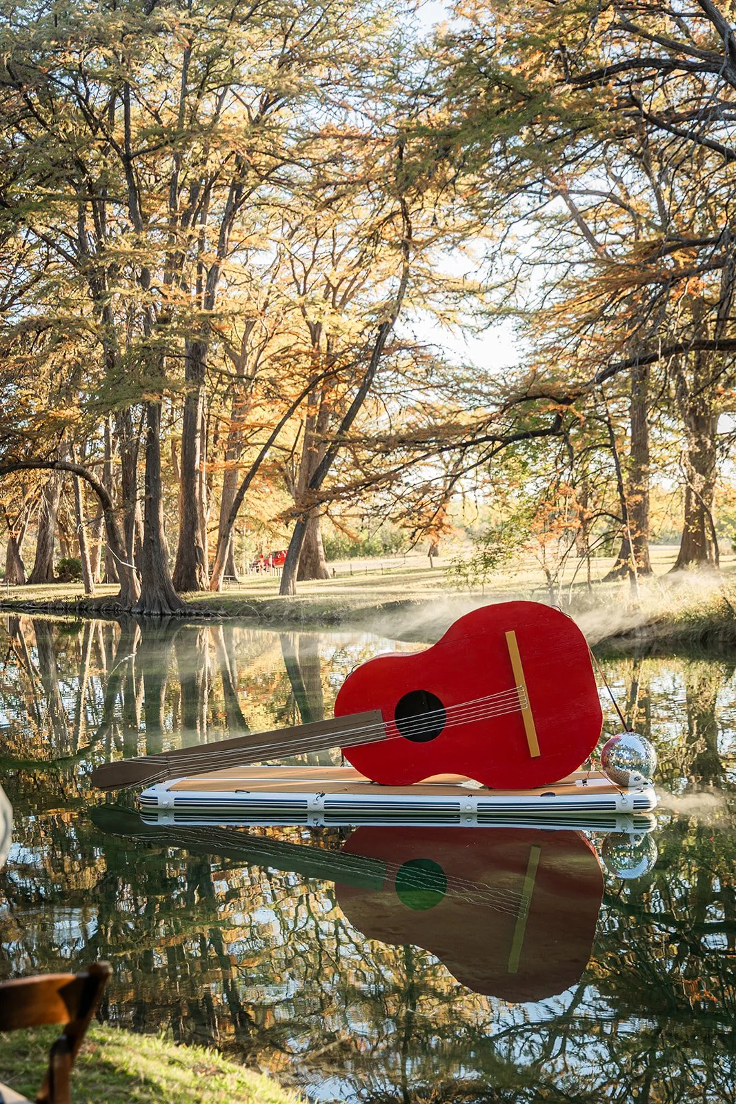 A small float in the shape of an acoustic guitar and a disco ball on a lake, with trees in fall colors and a clear sky in the background.