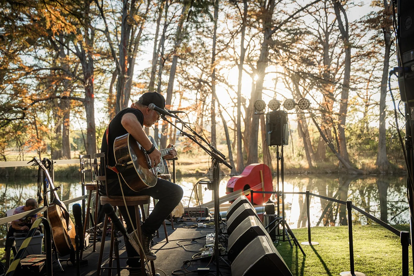 Musician playing guitar on stage outdoors near a lake, with trees and sunlight in the background during what appears to be late afternoon or early evening.