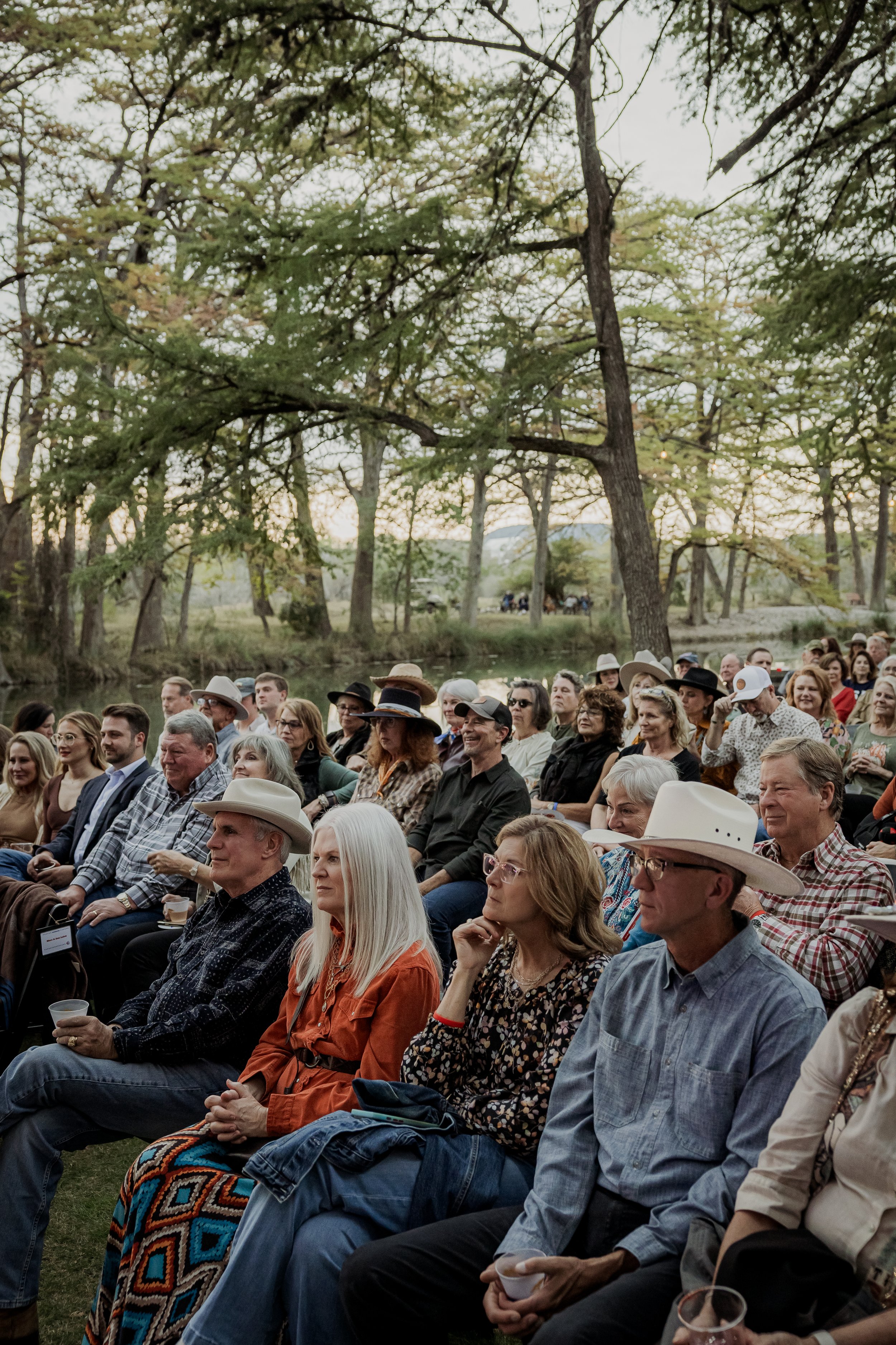 Crowd of people seated outdoors during a gathering or event near a body of water, surrounded by trees