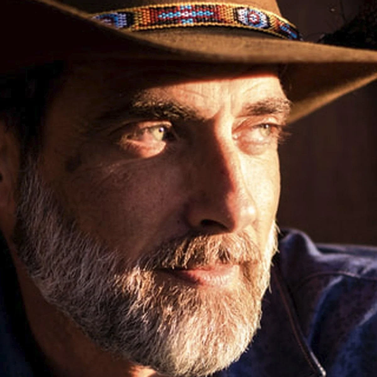 Close-up of a man with a beard wearing a cowboy hat with a colorful band, looking into the distance with warm sunlight on his face.