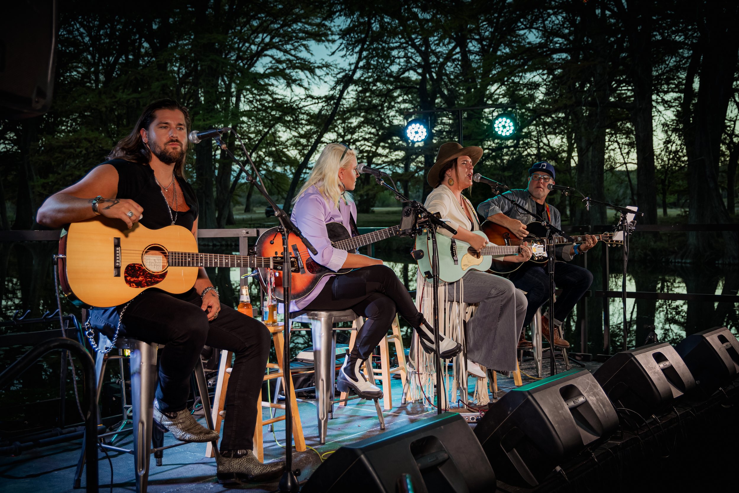 Four musicians performing on an outdoor stage during the evening, playing acoustic guitars and singing into microphones, with trees in the background and stage lights overhead.
