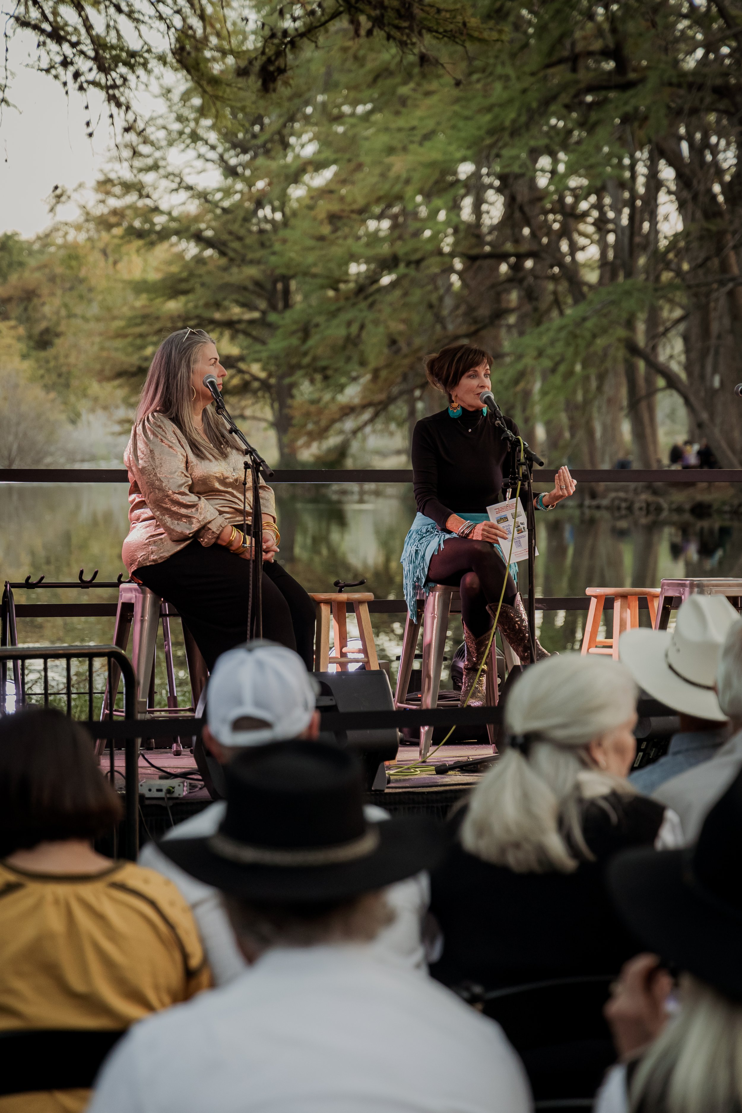 Two women sitting on stools on a stage outdoors near a body of water, speaking into microphones during a performance. An audience watches, some wearing hats. Trees are visible in the background.