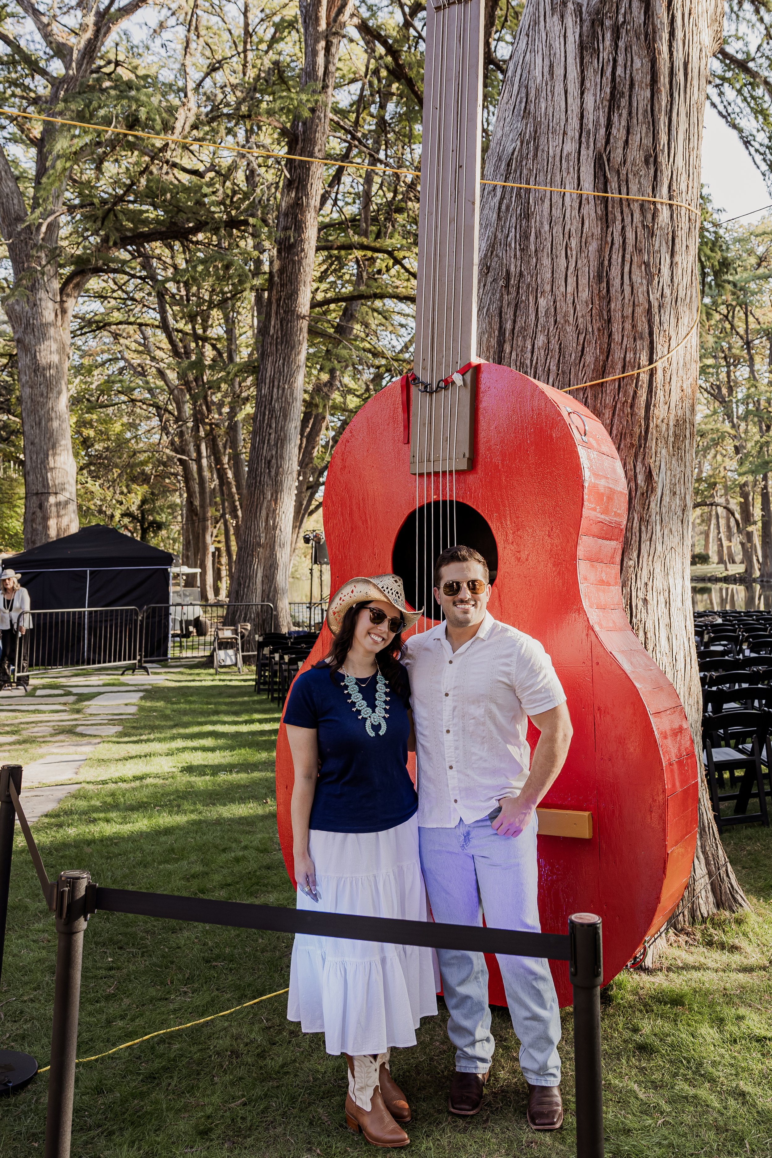 A smiling man and woman standing in front of a giant red guitar-shaped sculpture, with trees and chairs in the background.