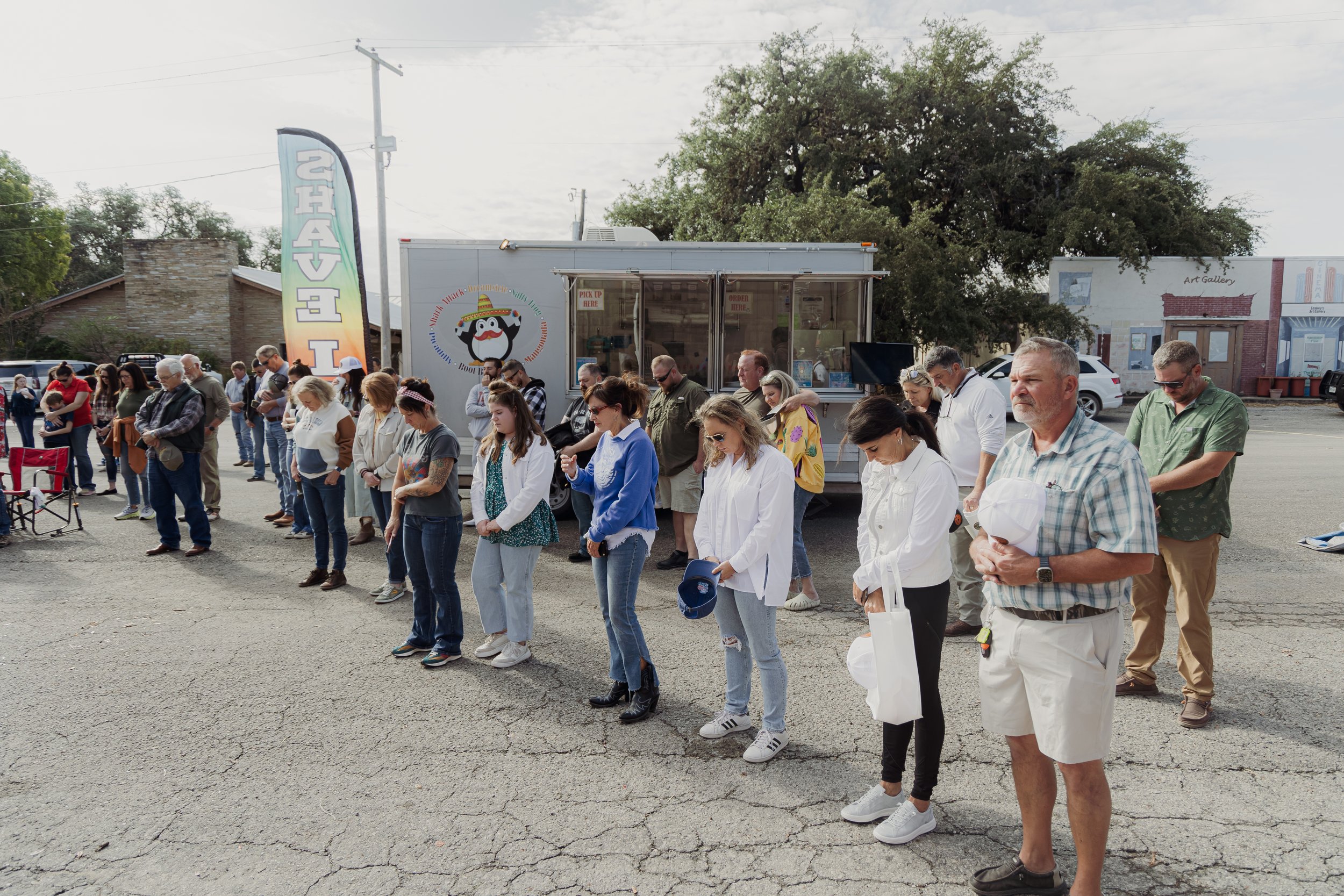A group of people standing in a line outdoors, with some holding white hats or bags, in a parking lot near a food truck, under a cloudy sky.