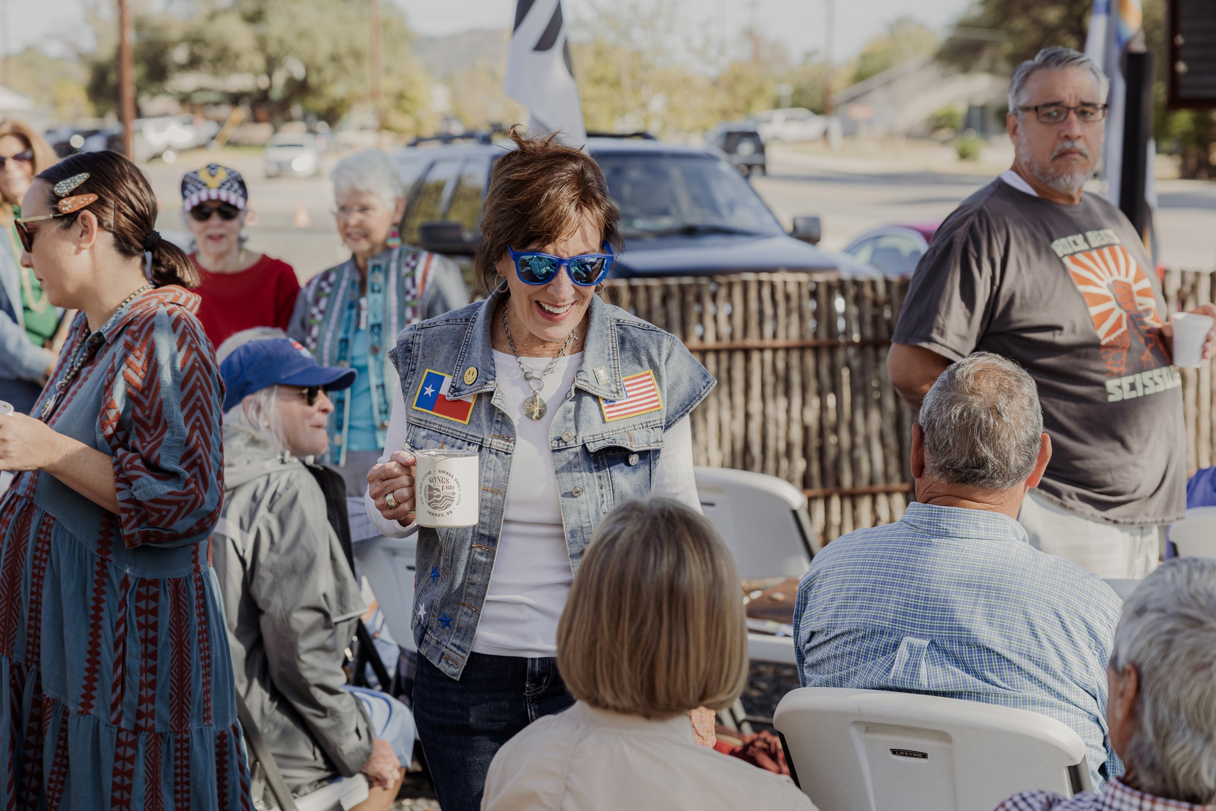 A group of people enjoying an outdoor gathering, with a woman in sunglasses and a denim vest smiling and holding a mug, surrounded by others seated and standing, in a setting with cars and trees in the background.