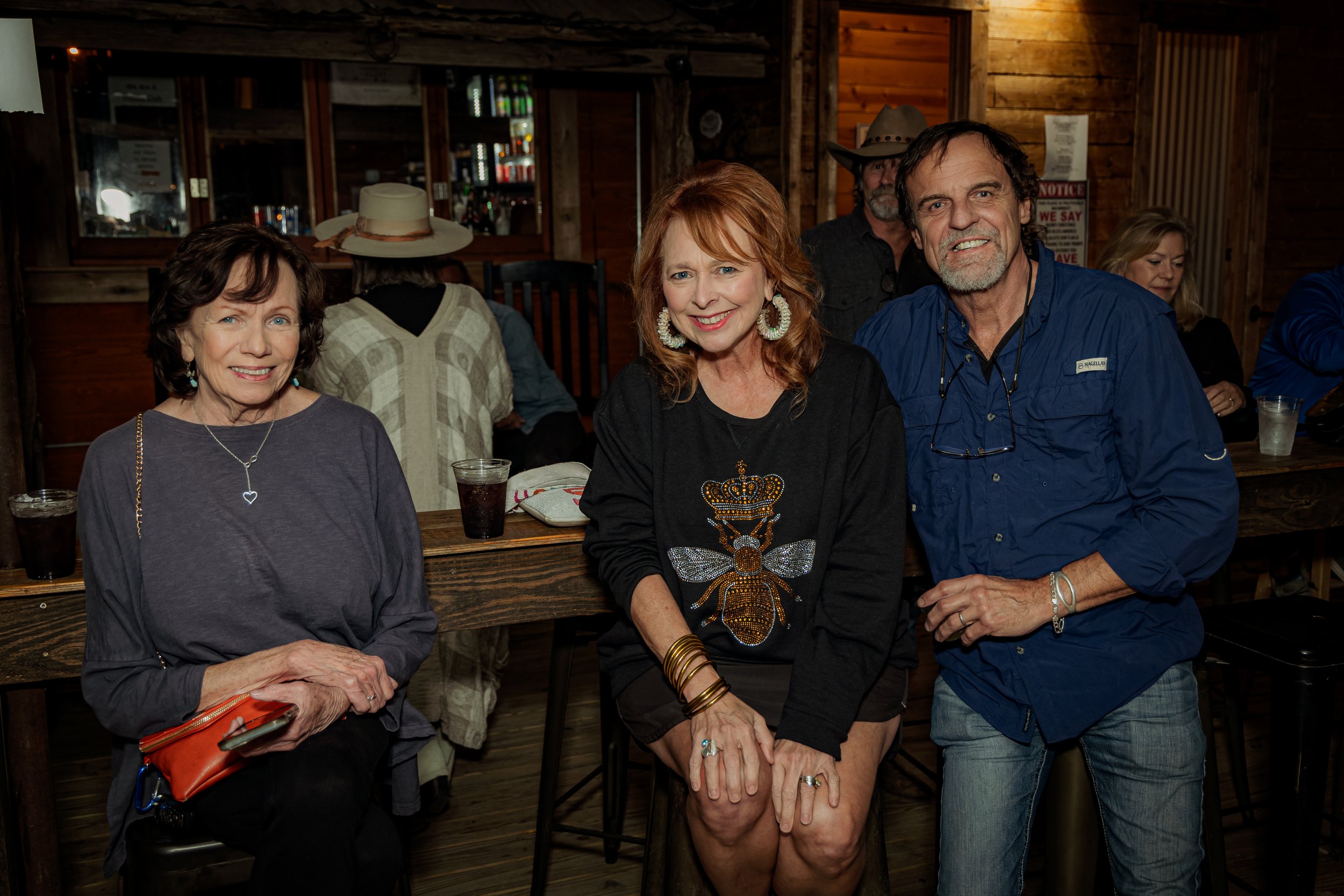 Three adults, two women and a man, sitting at a bar in a rustic wooden interior, smiling at the camera. Behind them are other patrons and a bar area with drinks.