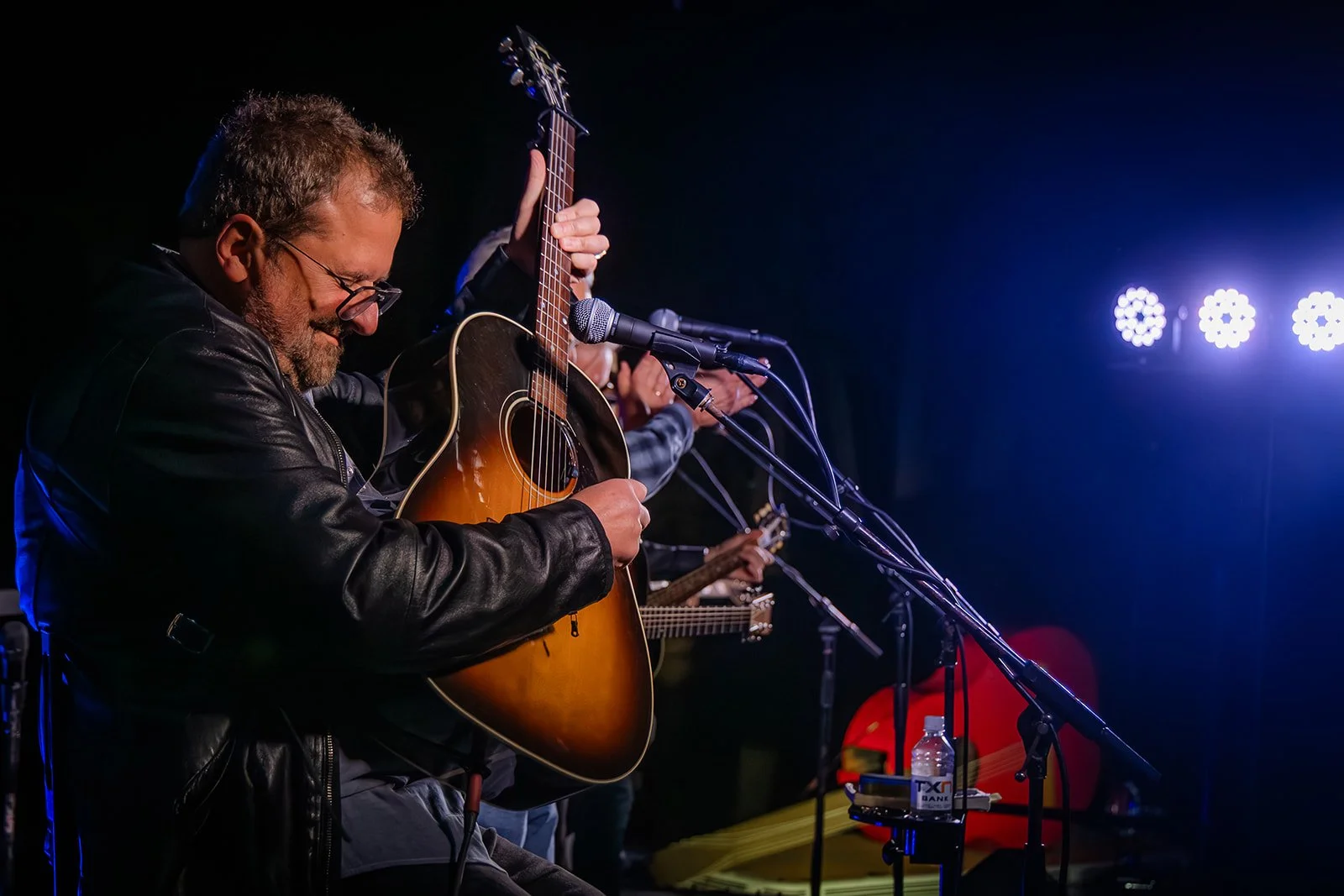 A man with glasses and a leather jacket playing an acoustic guitar on stage during a performance with microphones and stage lights.