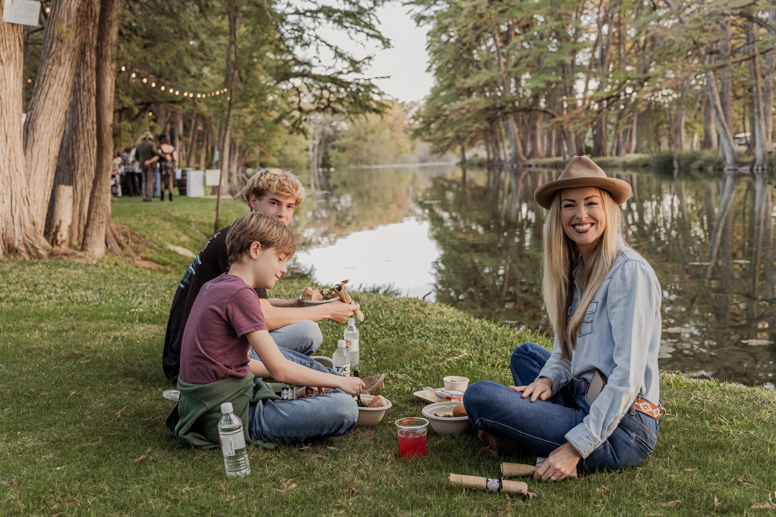 Family having a picnic on the grass by a lake with trees in the background and string lights overhead.