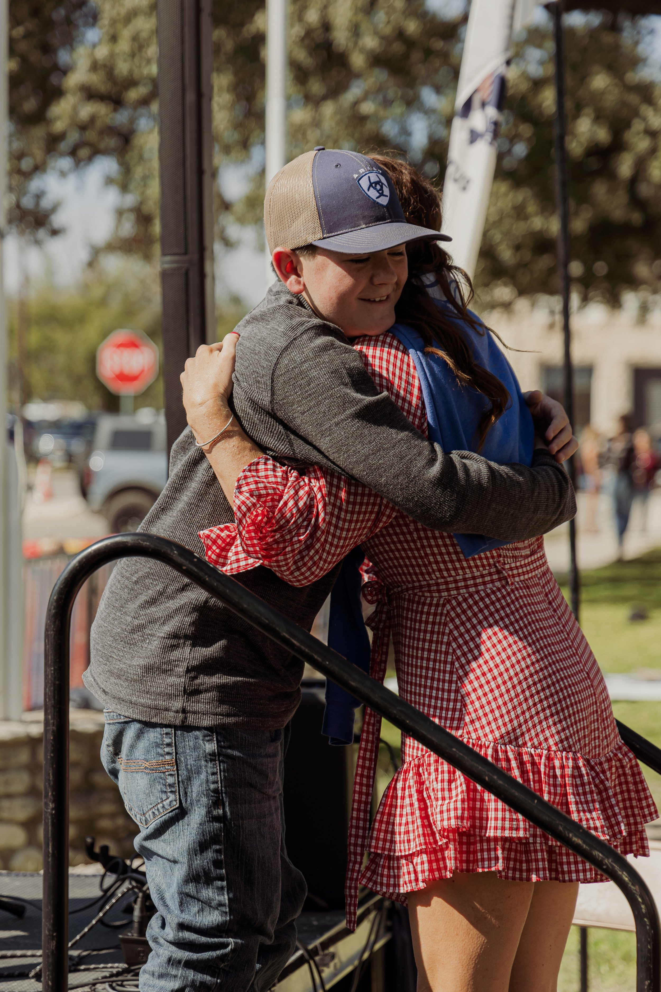 A young boy and girl hug each other on a stage outdoors. The boy is wearing a baseball cap and a grey sweater, and the girl is wearing a red and white checkered dress with a ruffled hem. There are trees and a stop sign in the background.