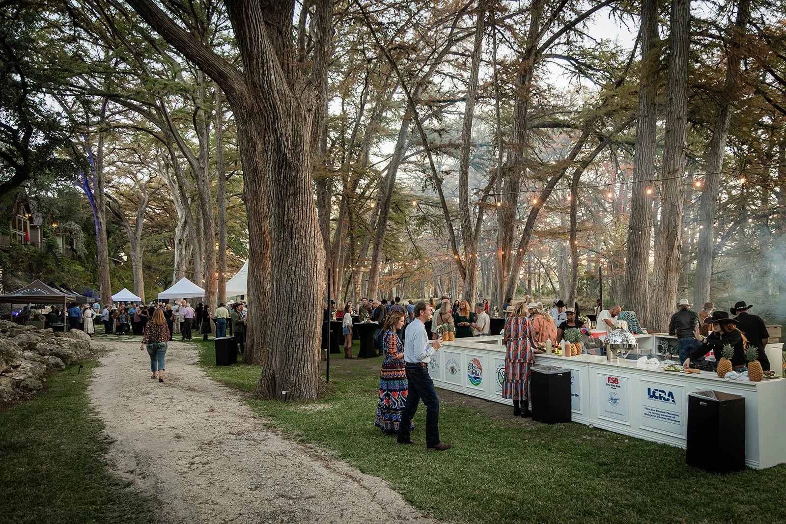 People gather at outdoor food and drink booths in a wooded park for a festival or event, with string lights hanging between trees and white tents in the background.