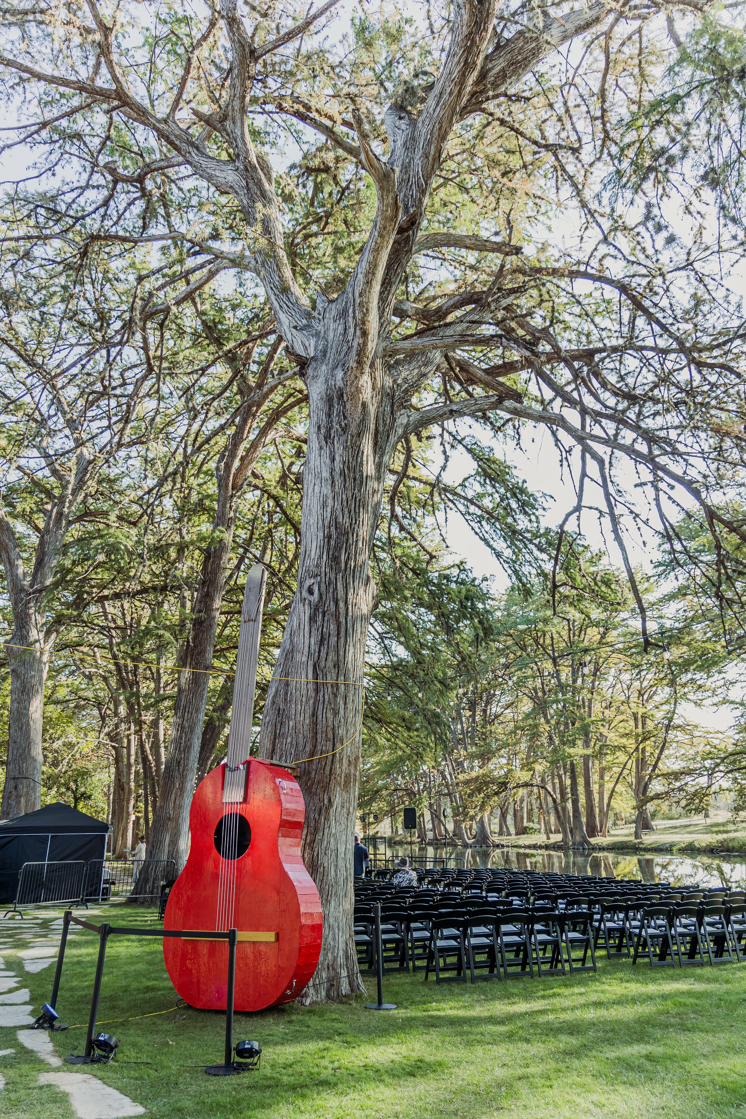 Outdoor event setup near a large tree with a red guitar sculpture, rows of black chairs, and a lake in the background.