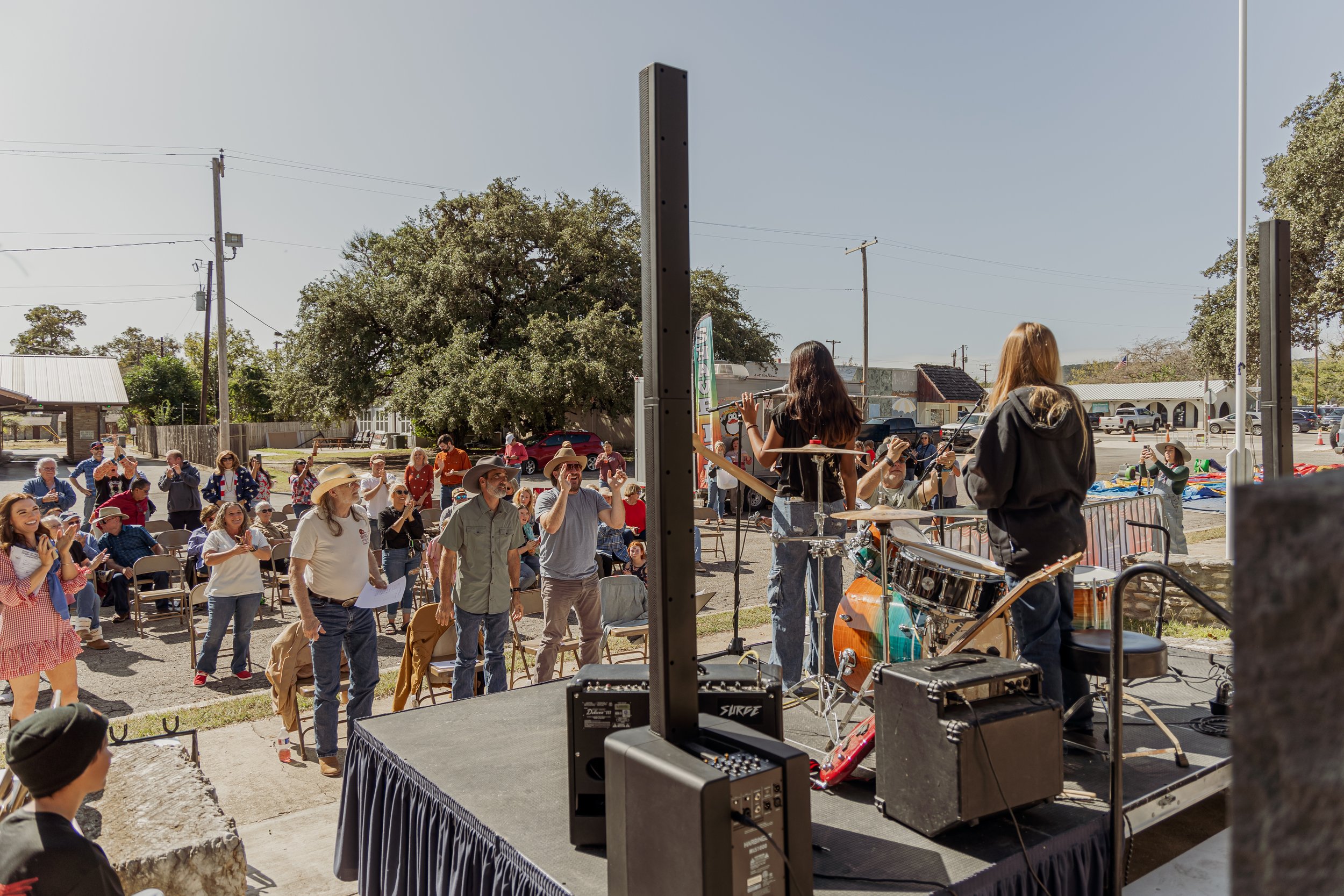 A live outdoor concert with an audience watching a band perform on stage during daytime, with people standing, clapping, and socializing.
