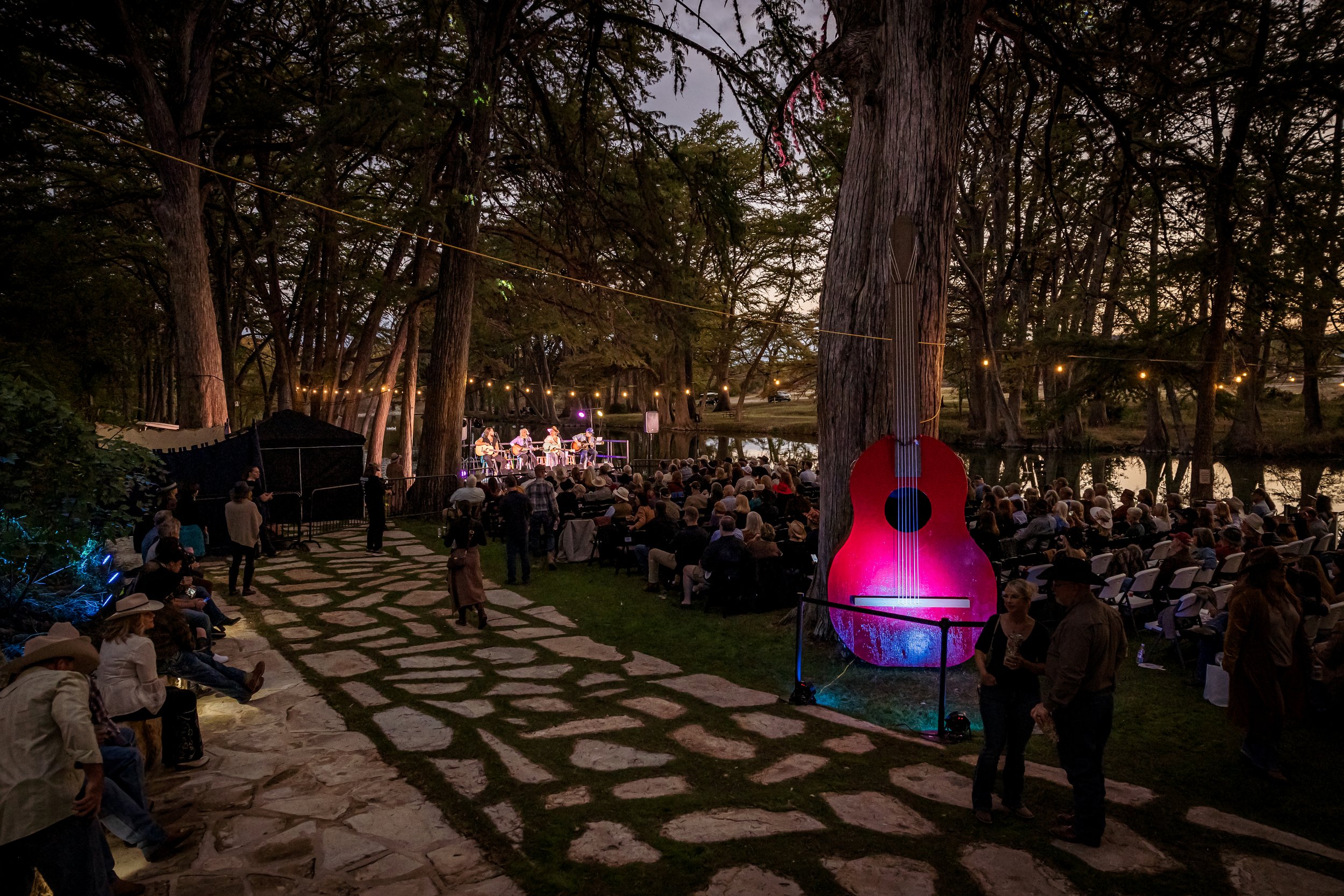 An outdoor concert at dusk with a stage of musicians to the left, audience seated and standing, and a large illuminated pink guitar sculpture on a tree to the right.