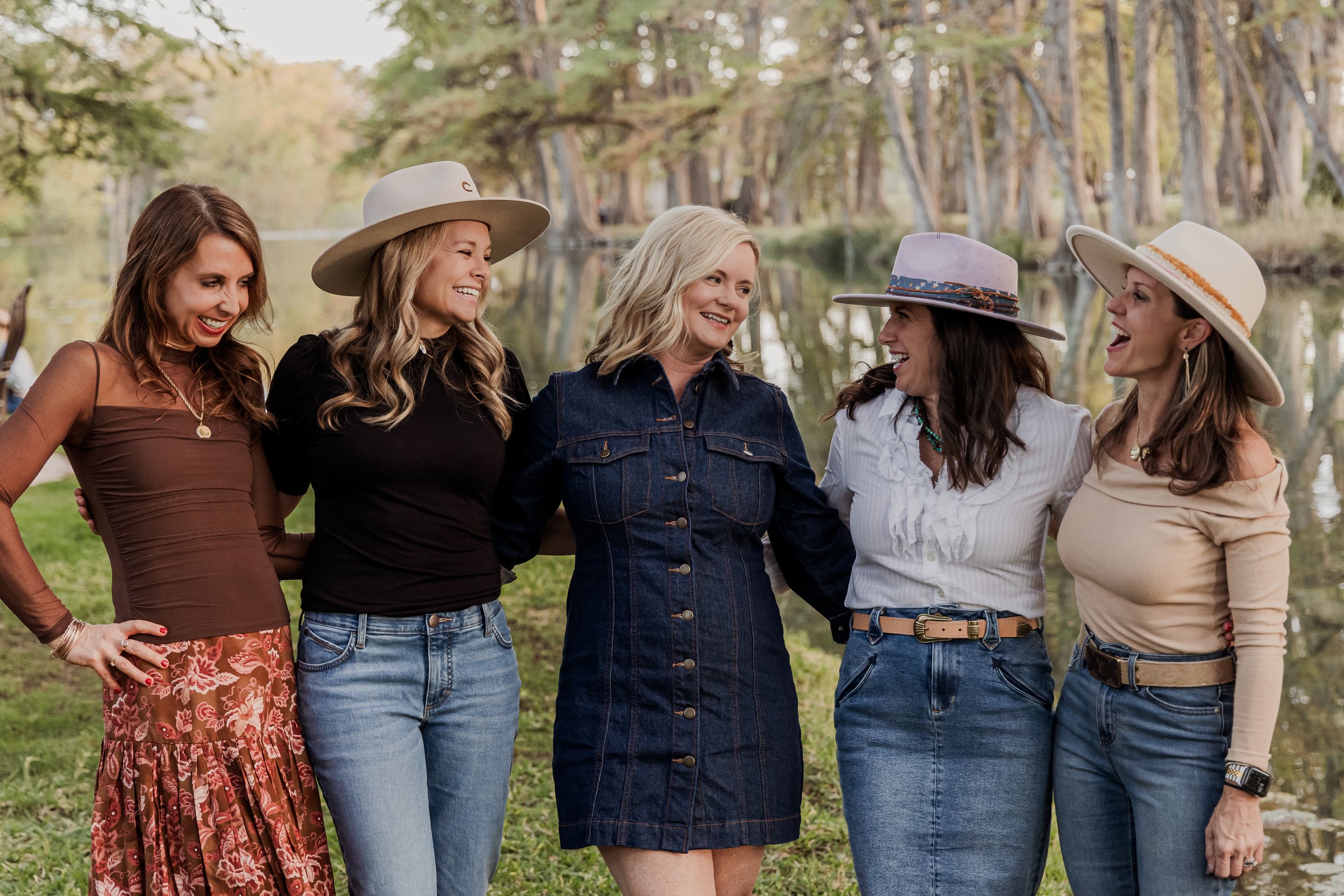 Five women standing arm-in-arm outdoors near a lake, smiling and laughing in a park with trees in the background.