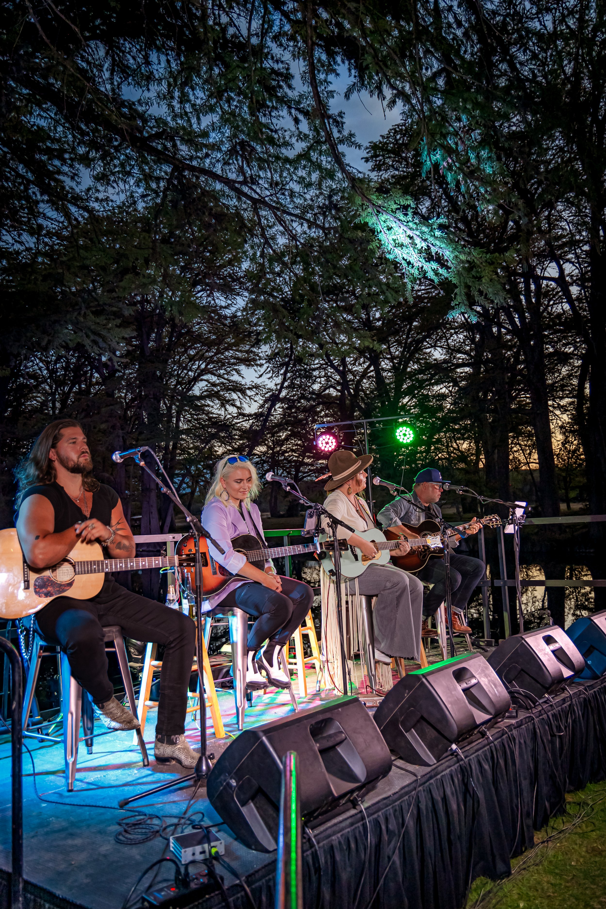 A musical band performing on stage outdoors during sunset, surrounded by trees, with stage lights shining on them.