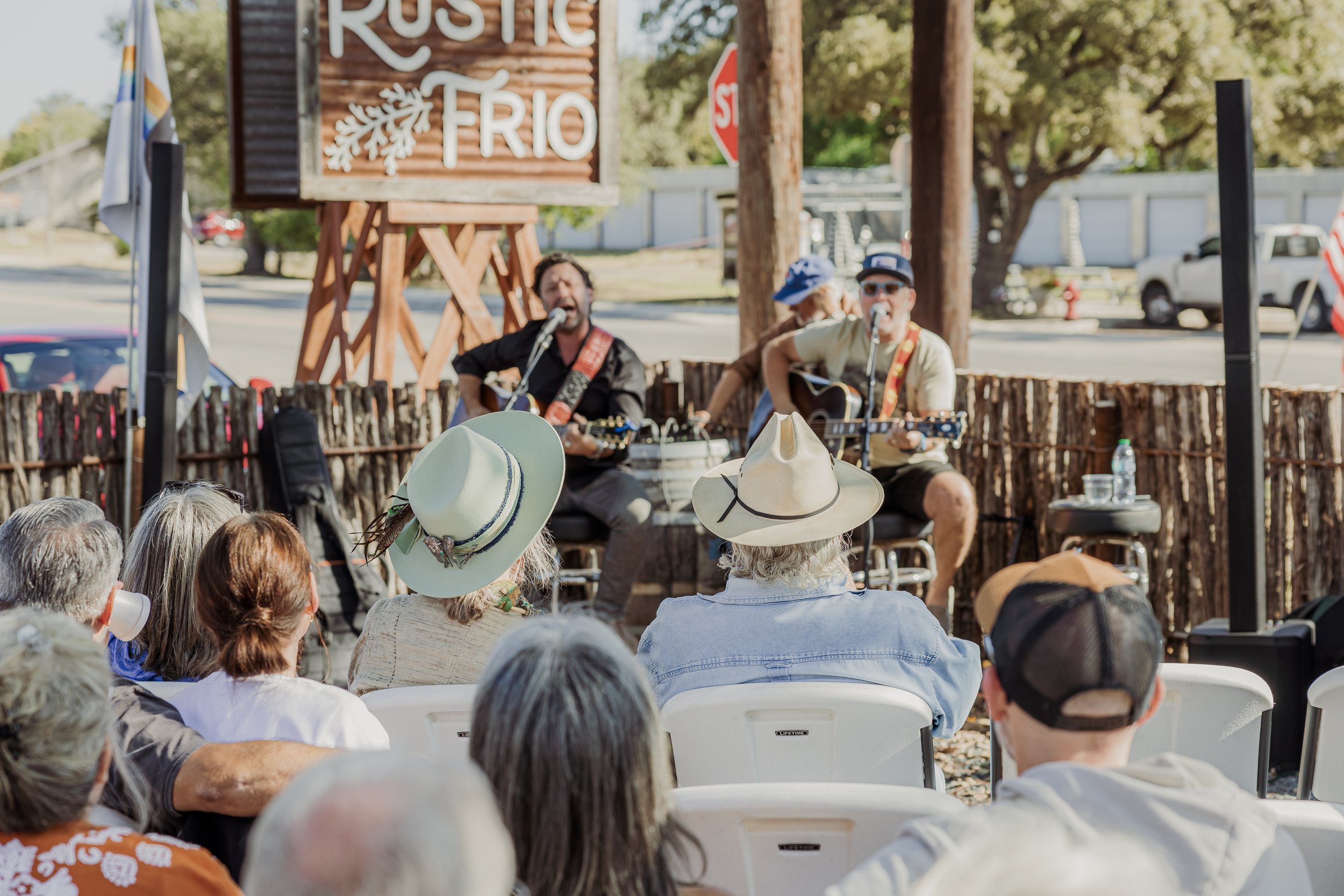 Musicians performing live outdoors for an audience at Rustic Frio, with audience members wearing wide-brimmed hats and sitting on white chairs.