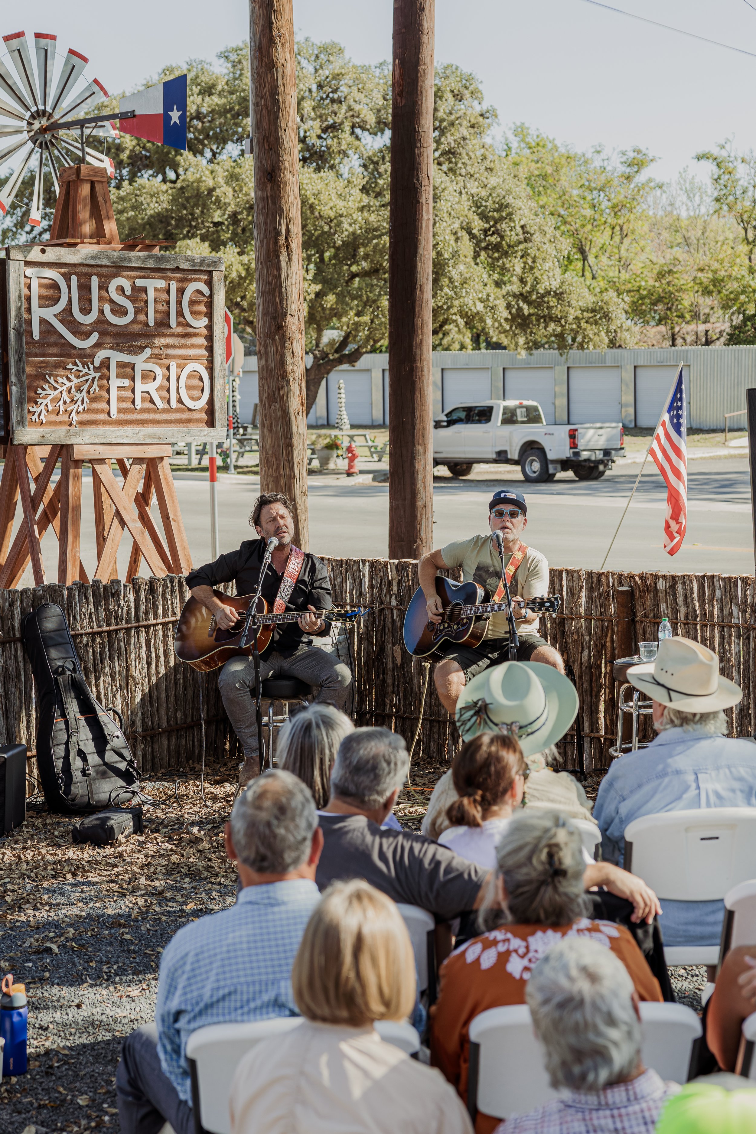 Two musicians playing guitars and singing during an outdoor event at Rustic Rio with an audience seated on white chairs, American flags, and a sign reading 'Rustic Rio'. The setting is rustic with trees, a pickup truck in the background, and a windmi