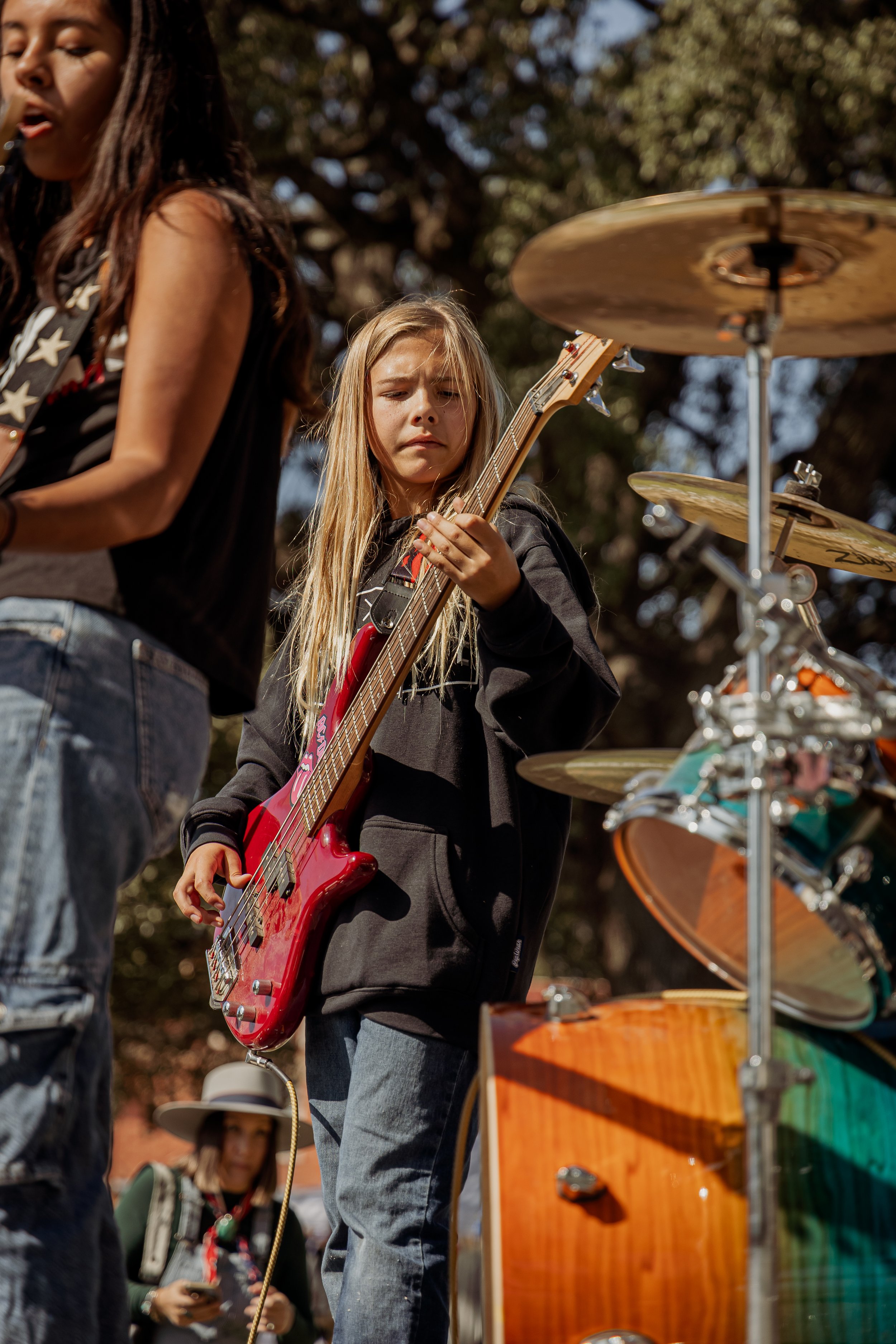 A young girl playing a red electric bass guitar outdoors, surrounded by percussion instruments and other musicians, with trees in the background.