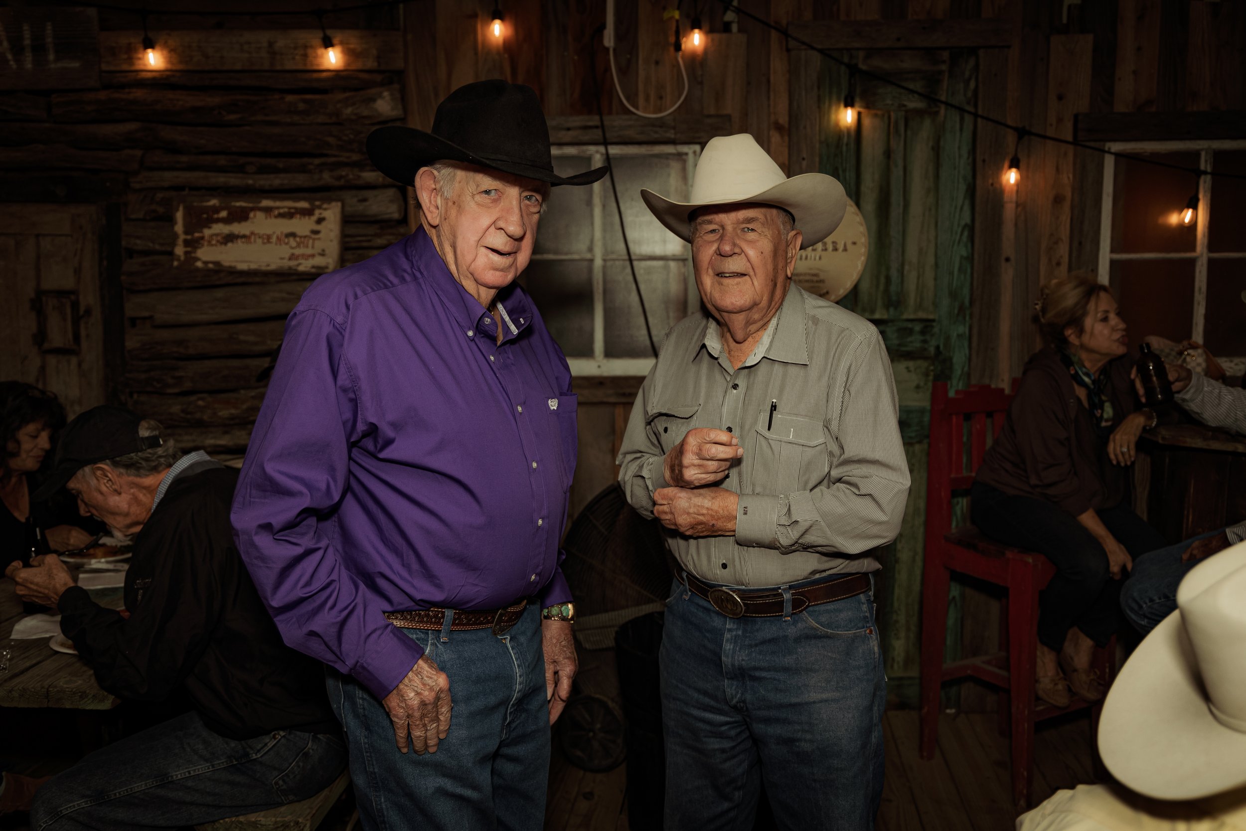 Two elderly men in cowboy hats and Western shirts standing together inside a rustic wooden building, with other people sitting at tables in the background.