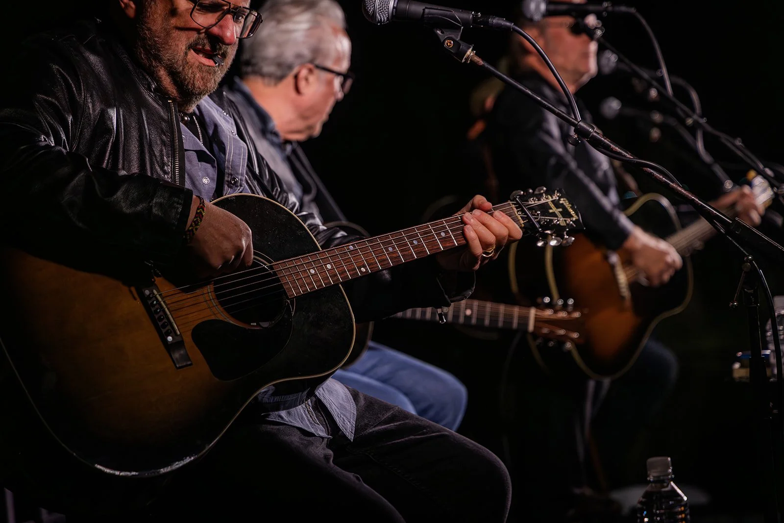 A group of musicians playing acoustic guitars during a live performance, with microphones set up in front of them.