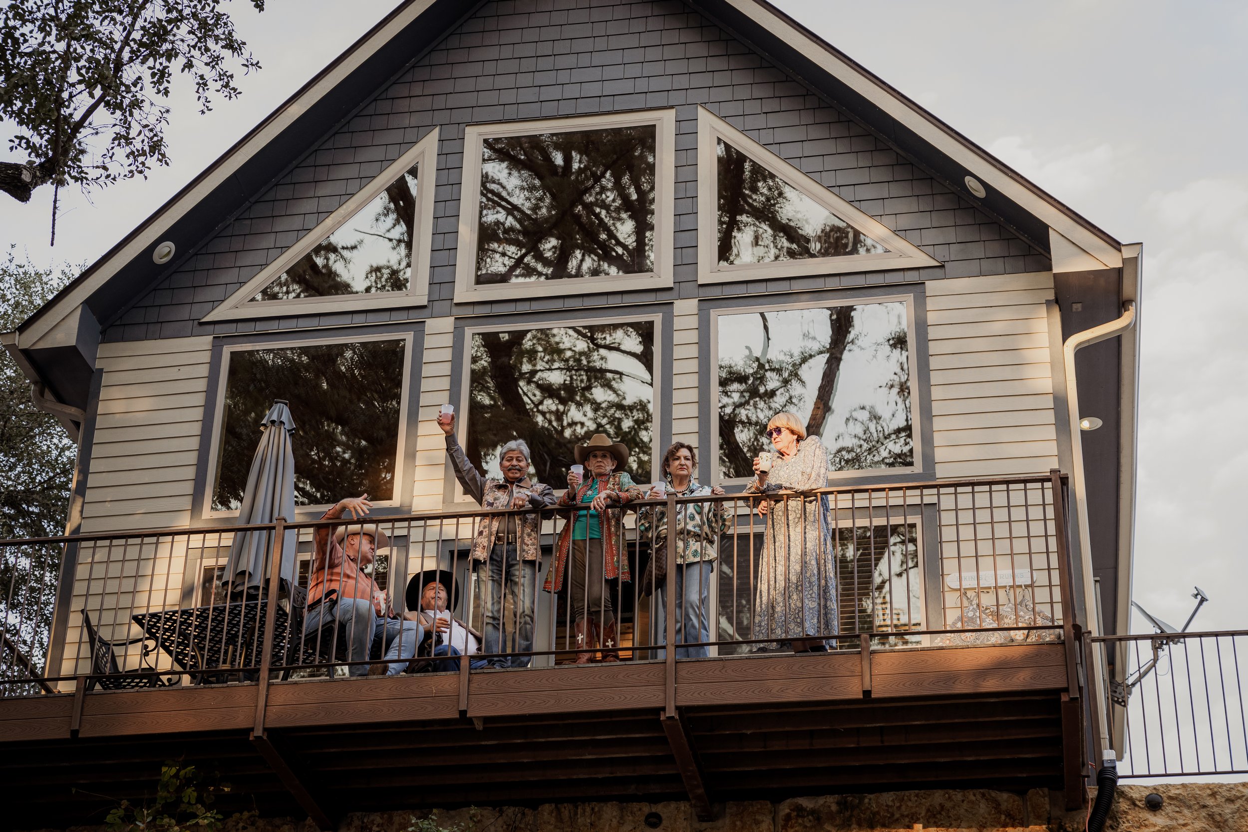 Group of six people standing on a balcony in front of a house with large mirrored windows, some holding drinks, and one person raising a glass for a toast.