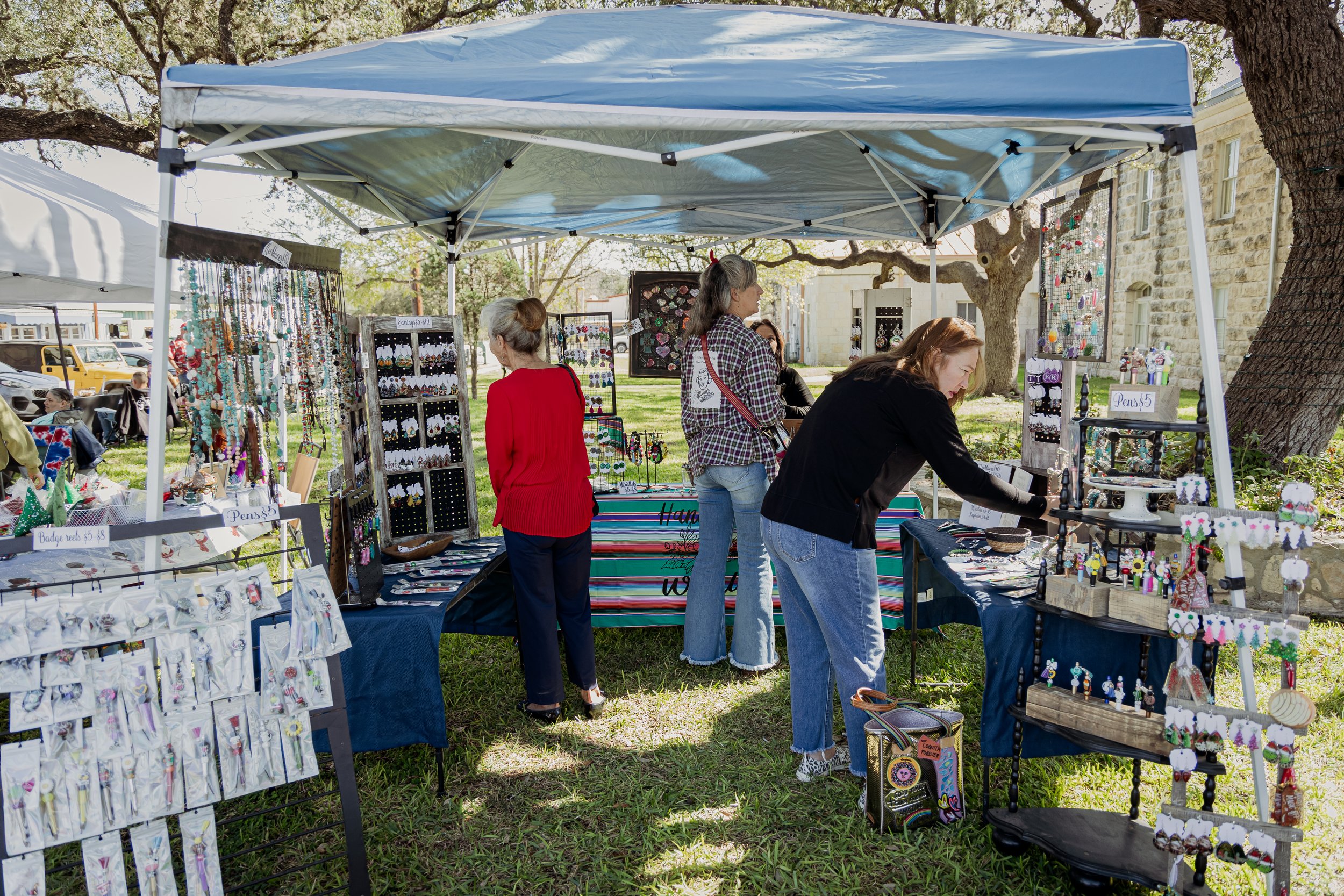People browsing jewelry and crafts at an outdoor market stall under a blue canopy, with trees and stone buildings in the background.