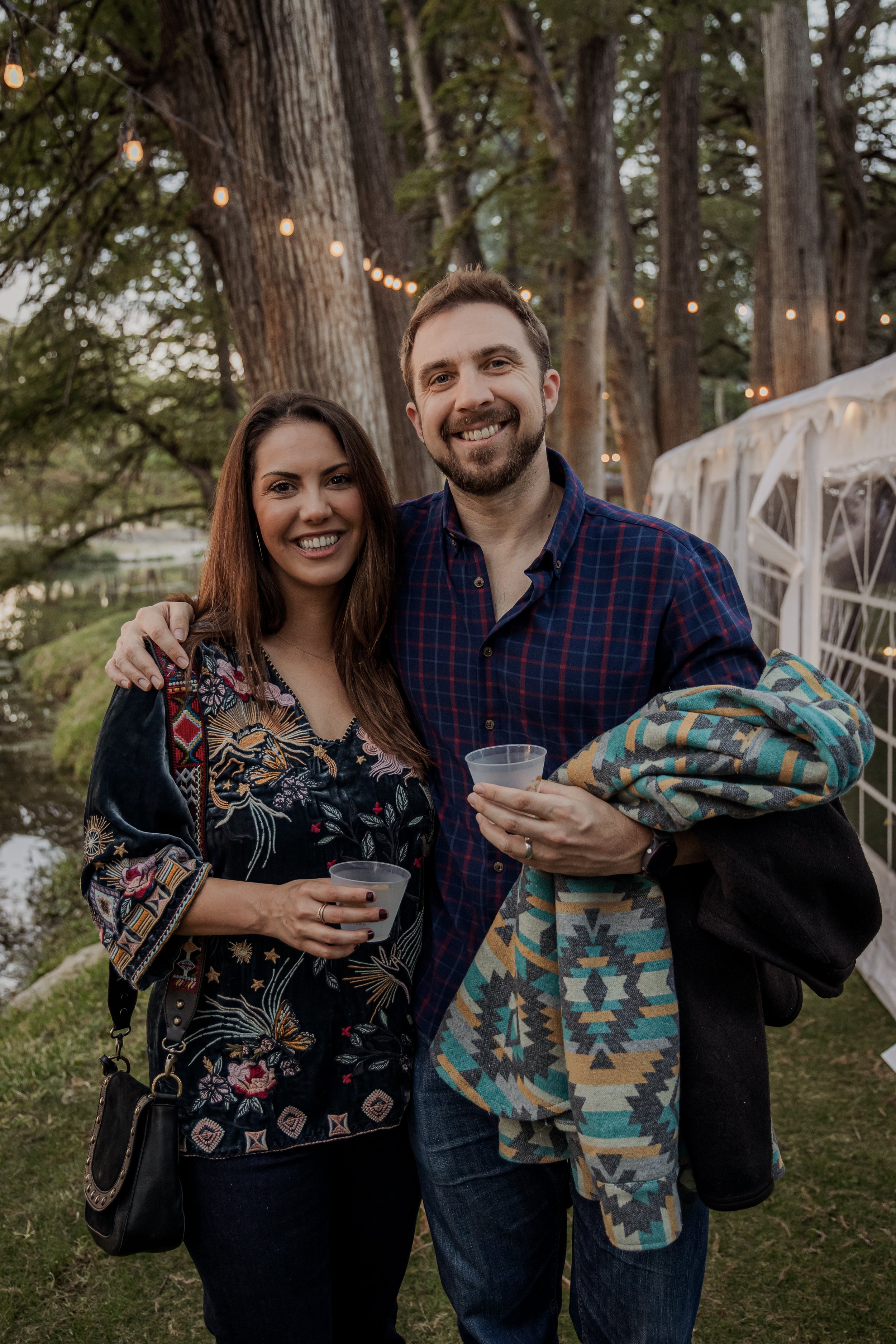 A woman and a man standing outdoors at dusk, smiling and holding drinks, with string lights and large trees in the background.
