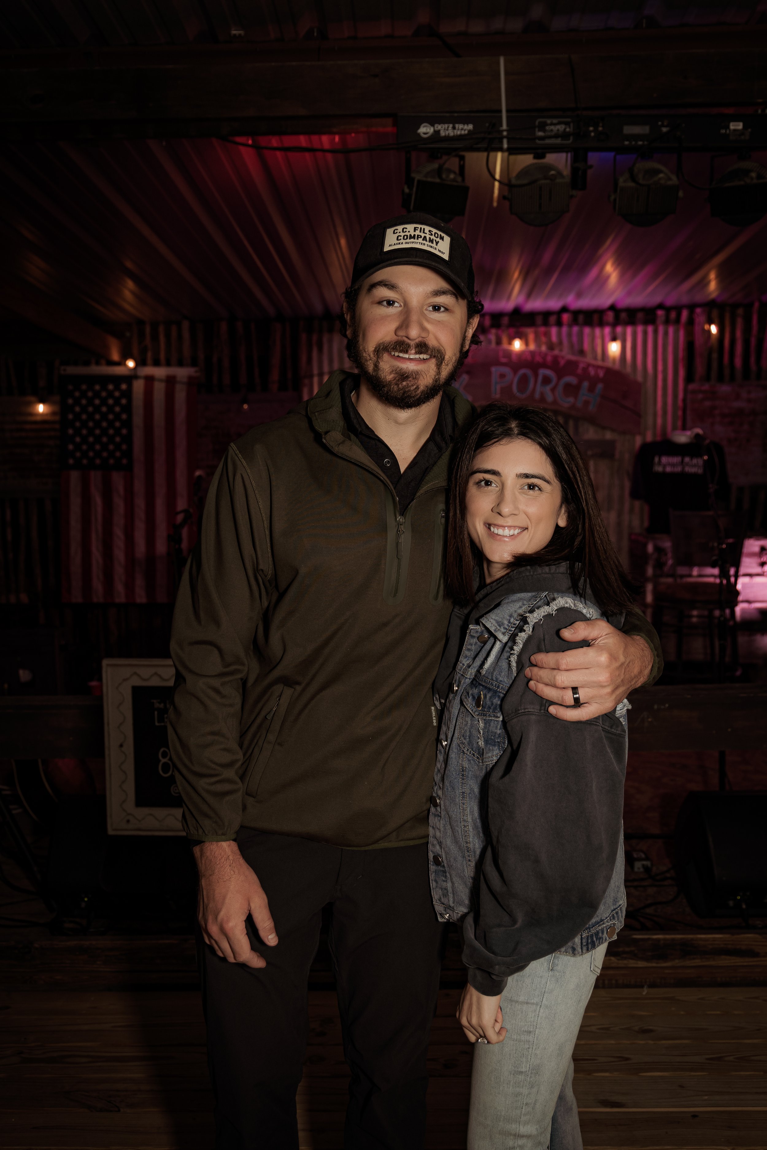 A man and woman standing together, smiling and hugging in a dimly lit venue with a stage and American flag in the background.