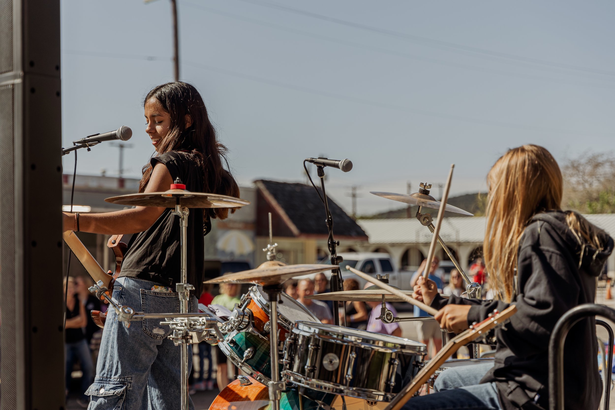 Two young girls performing music outdoors, one playing drums and the other singing into a microphone, with a crowd and buildings in the background.